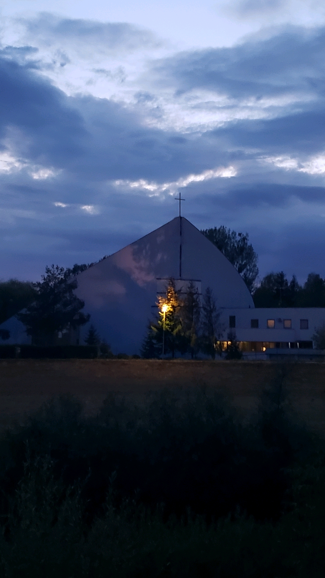 The image depicts a modern church building at dusk, situated in Prešov, Slovakia. The sky is filled with dramatic, dark clouds, with hints of light breaking through. A prominent cross stands atop the triangular roof of the church. The building itself is white with a distinctive curved facade. To the right of the main structure, a lower, rectangular section of the building features several illuminated windows. In front of the church, a single streetlamp casts a warm glow on a small evergreen tree and surrounding foliage. The foreground is dominated by a dense, dark area of vegetation, possibly bushes or trees, obscuring the ground. The overall atmosphere is serene and slightly melancholic, indicative of twilight.