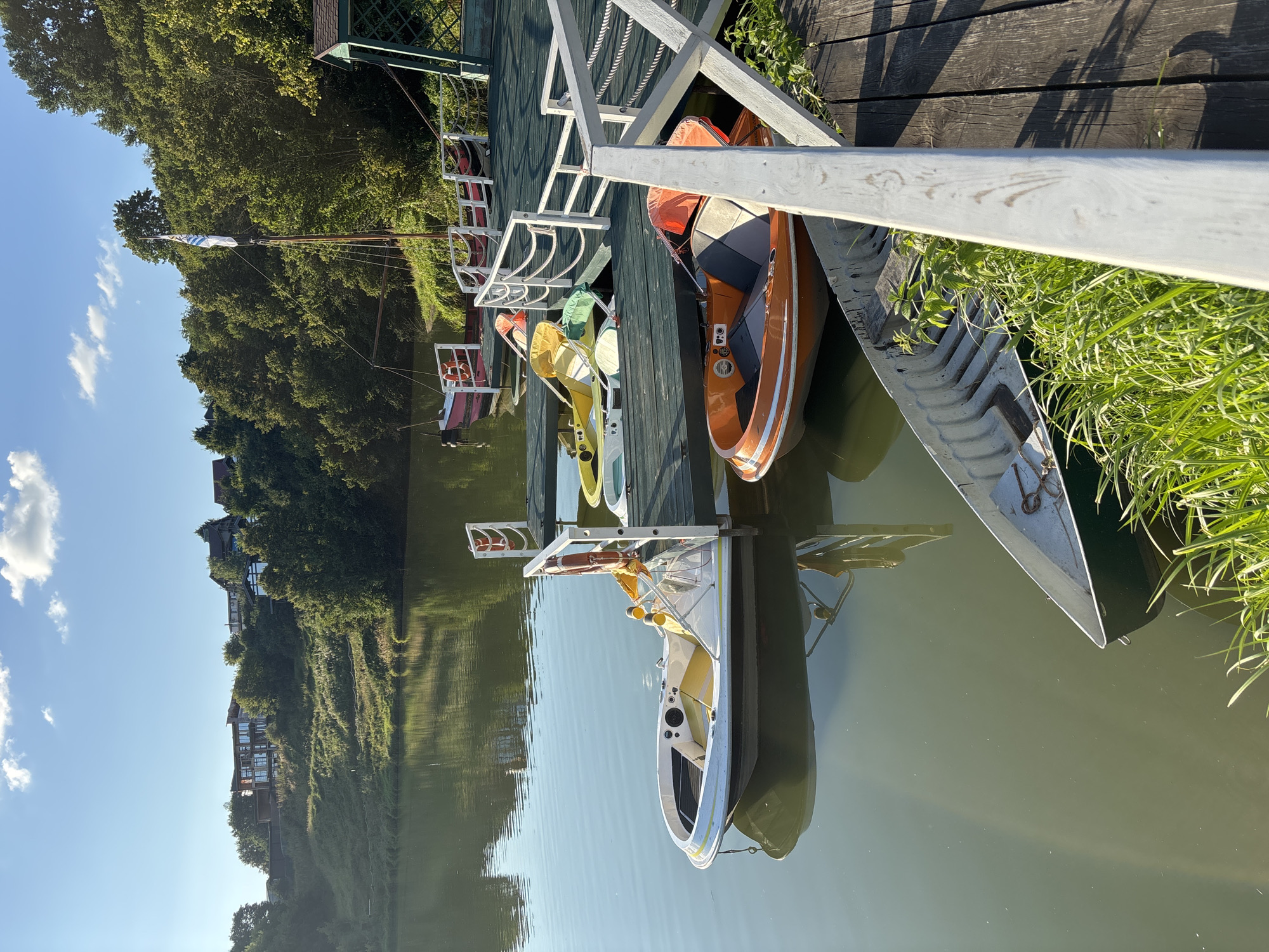 The image depicts a serene waterfront scene at the Romashkovo marina, Russia. Several small boats, including a white and yellow pedal boat and an orange boat with grey seats, are docked at a wooden pier. The pier is adorned with white railings and ropes. In the background, lush green trees line the embankment, and a few houses are visible on the hilltop. The water is calm, reflecting the clear blue sky with scattered white clouds. A sailboat with a white and blue flag is anchored further out in the water. The overall atmosphere is peaceful and idyllic, suggesting a pleasant day for boating.