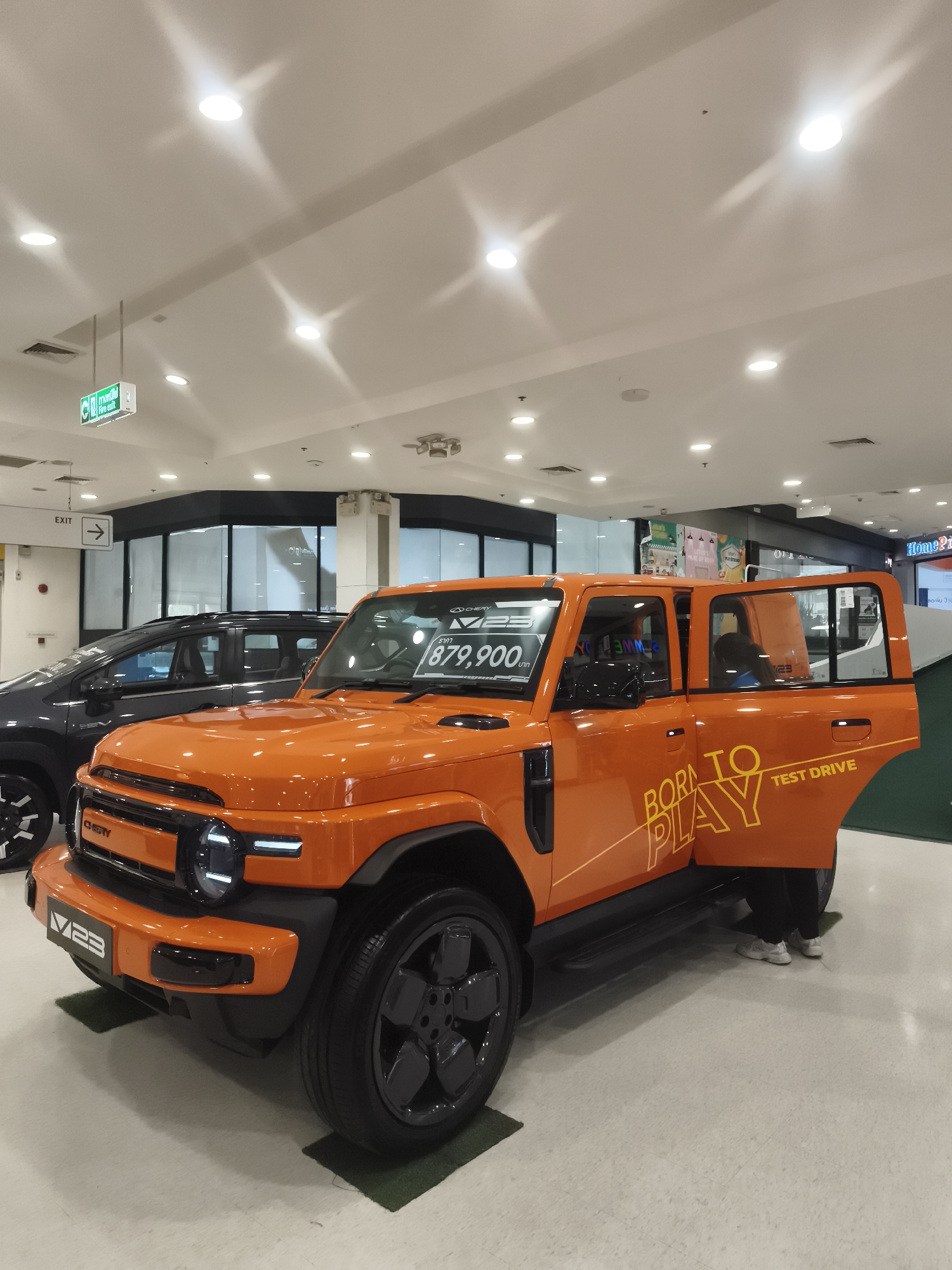 An orange Chery V23 SUV is parked on a light gray tiled floor in what appears to be an indoor showroom or mall in Ko Samui District, Thailand. The car is angled towards the viewer, with its driver-side door open. The phrase "BORN TO PLAY TEST DRIVE" is emblazoned in yellow on the side of the car. A price tag on the windshield reads "879,900". A dark-colored SUV is parked behind the orange vehicle. The ceiling features recessed lighting and ventilation grates. An "EXIT" sign with an arrow is visible on the left side of the frame, along with a green "FIRE EXIT" sign. Two individuals, wearing dark pants and white sneakers, are standing near the open door of the orange SUV. The overall lighting is bright and even, suggesting an indoor commercial space.