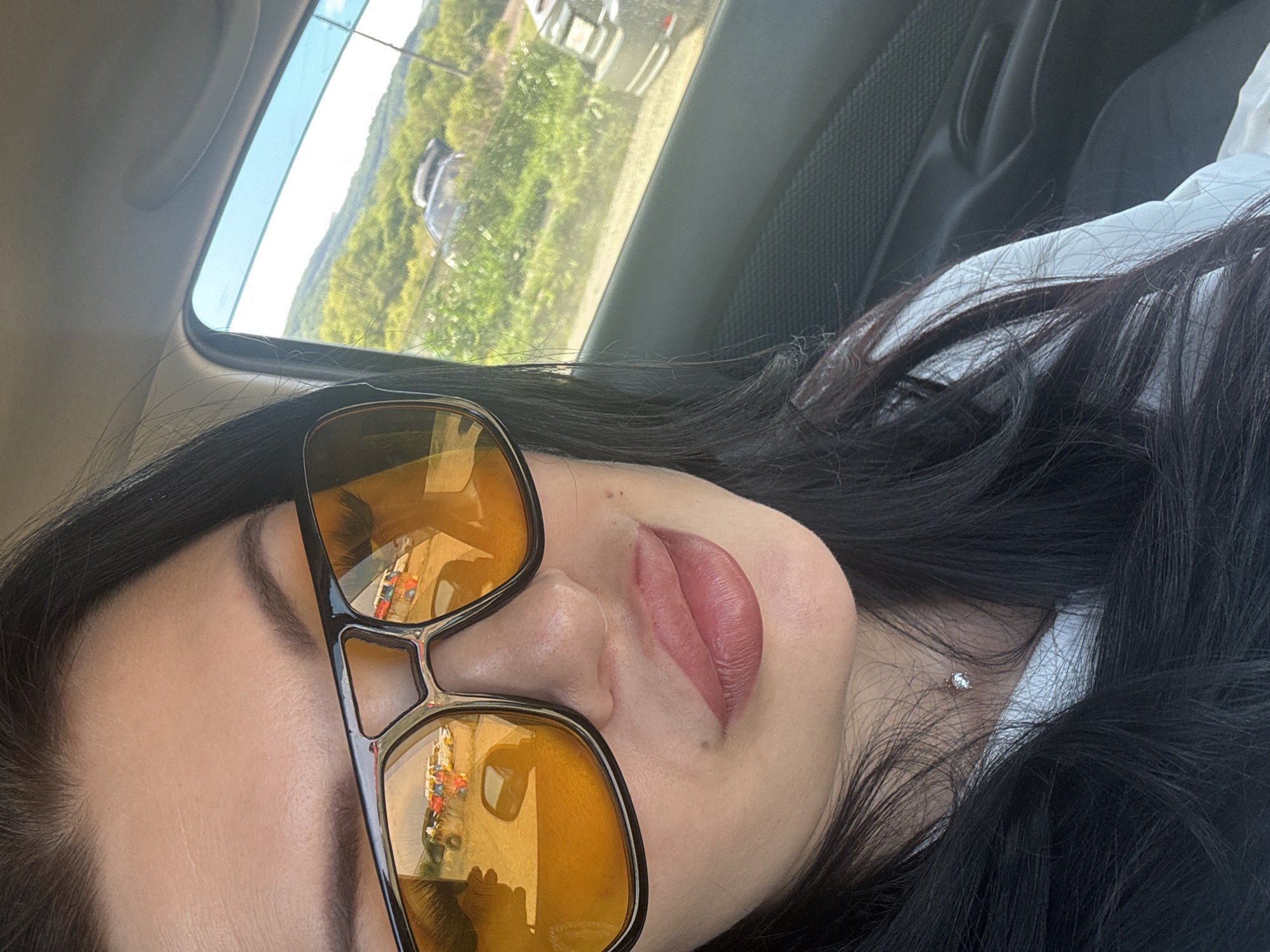 A woman with dark hair is pictured indoors, possibly in a car, wearing large amber-tinted sunglasses. Reflected in the lenses are figures and structures. The window behind her shows a green, possibly hilly landscape and a glimpse of another vehicle. Based on the reflections in the glasses, she is possibly located in Vladivostok, Russia.