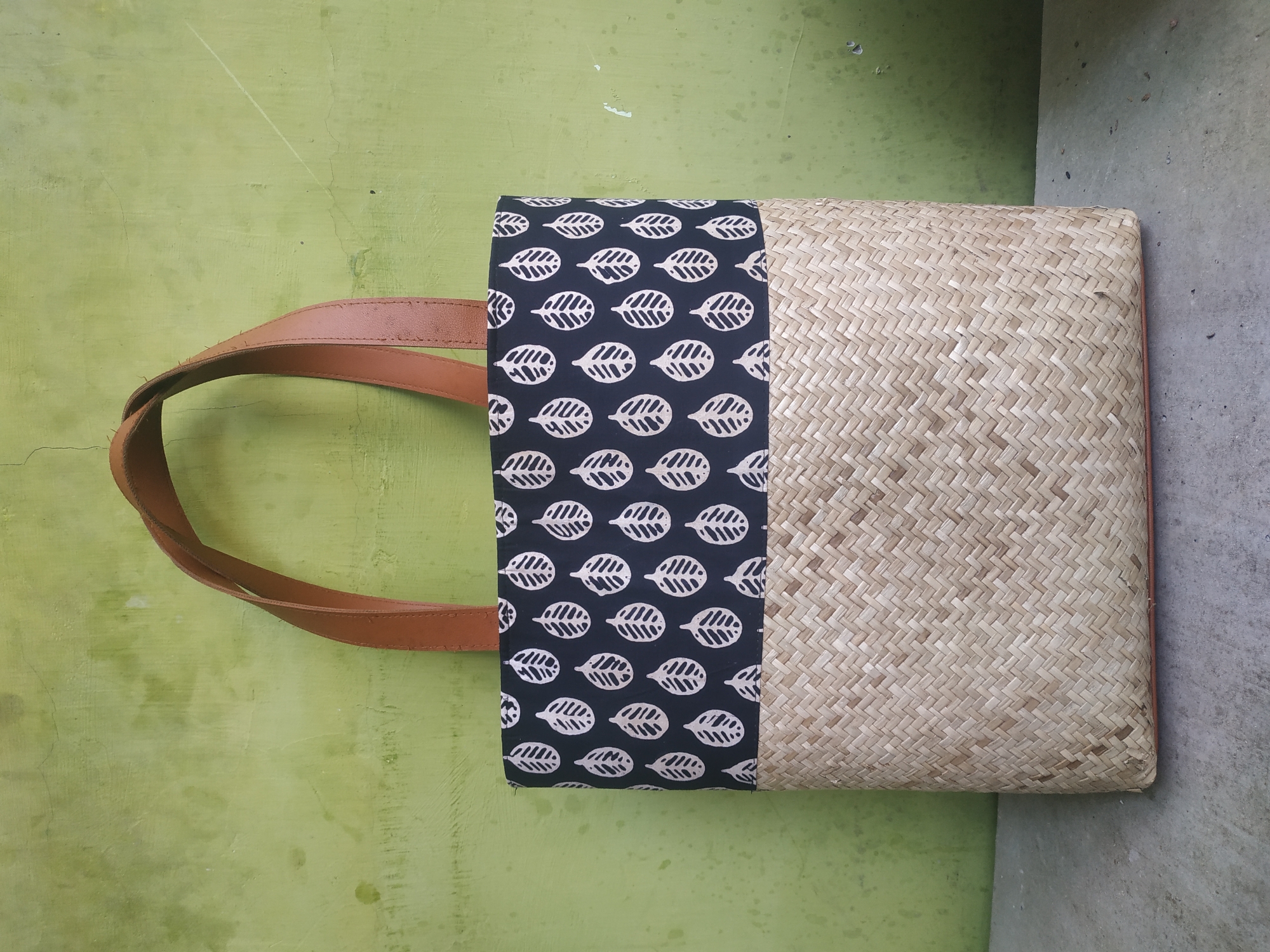 A handcrafted tote bag, featuring a lower section woven from natural straw-like material and an upper section of dark fabric with a repeating white leaf pattern, stands against a textured, weathered green wall. The bag has two brown leather straps for carrying. The scene is set on a concrete surface, suggesting an outdoor or semi-outdoor location. The image was captured in Jember, Indonesia.