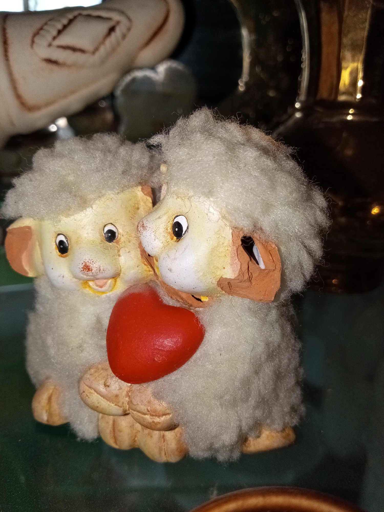 Two sheep figurines are shown in the foreground, holding a red heart. The sheep are cream colored with a fluffy exterior and painted faces. One is positioned slightly behind the other, with their heads touching. The background contains blurry shapes that appear to be other objects. There is no evidence to suggest that the location is Щучье Озеро, Russia.