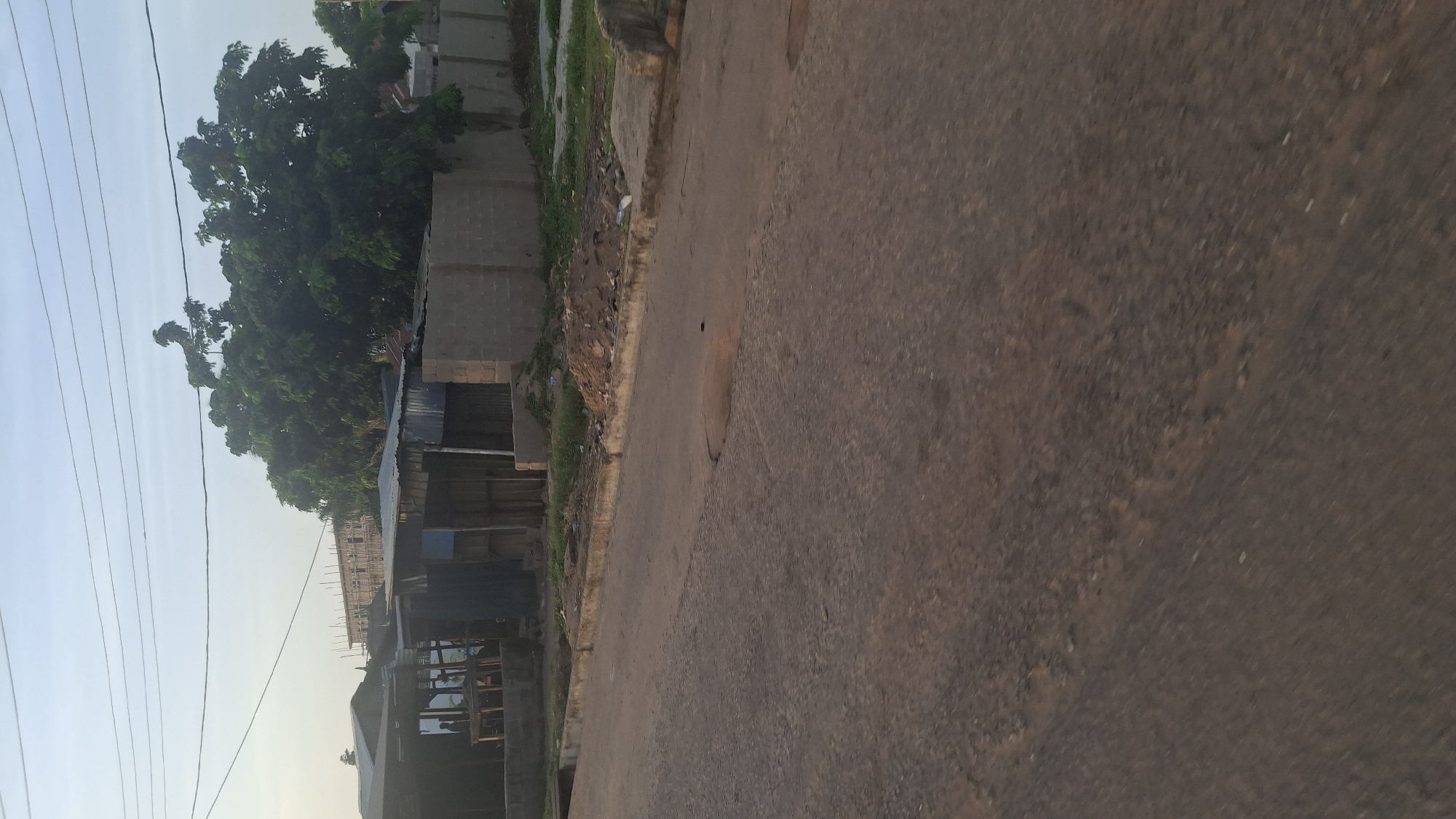 This media file depicts an outdoor scene in Jalingo, Nigeria, likely during the late afternoon due to the warm, golden light illuminating the landscape. The image is oriented vertically, with the road dominating the right side of the frame and the buildings and vegetation on the left.
The scene appears to be a residential or developing area. On the left, a dense canopy of lush green trees stands tall, suggesting a warm climate. Beneath the trees, a row of structures, some appearing to be under construction with scaffolding, and others with corrugated metal roofing, are visible. These buildings are made of concrete blocks. In the foreground of the left side, a complex of what looks like small shops or workshops with wooden frames and corrugated metal roofs are lined up. Overhead, a network of power lines stretches across the sky, indicating the presence of electricity infrastructure.
The road in the foreground is made of a reddish-brown, packed earth or asphalt material. It appears to be a well-traveled path. A circular manhole cover is visible on the road surface. The edge of the road is lined with dirt, some scattered debris, and patches of grass, indicating a somewhat unkempt but lived-in environment.
There are no people visible in the frame. The overall mood is tranquil, capturing a slice of daily life in a Nigerian town. The lighting suggests a time when the sun is lower in the sky, casting soft shadows. The weather appears clear and pleasant. No text is visible in the image.