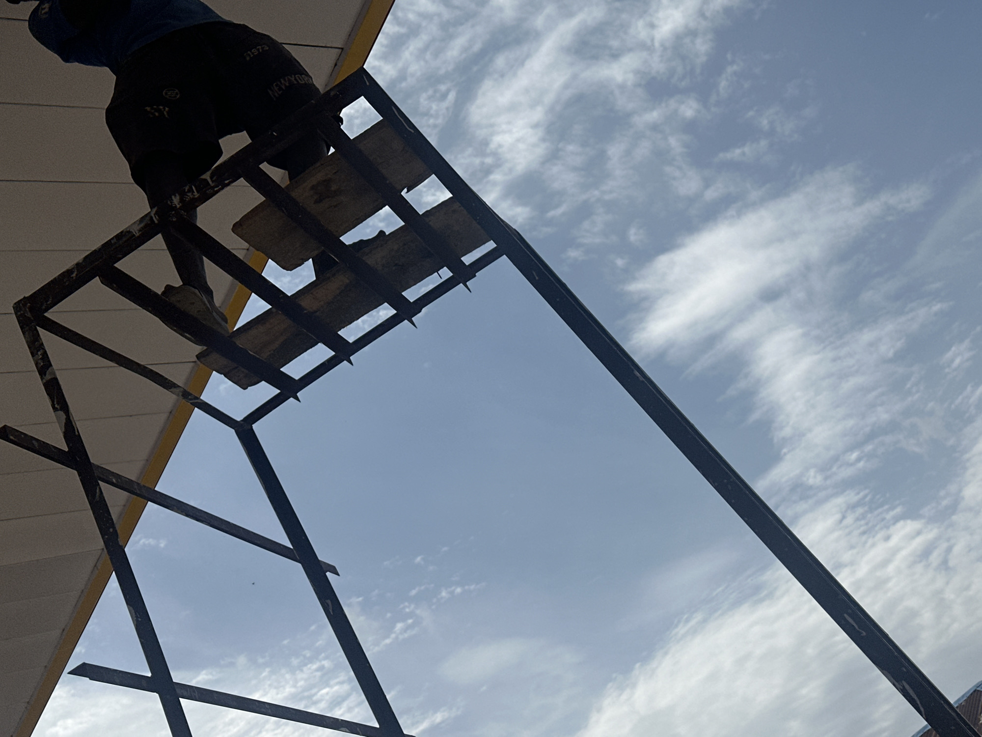The image shows a low-angle shot looking up at a structure against a bright, cloudy sky. The structure appears to be made of dark metal beams, forming a lattice-like framework. Within the framework, there are several wooden planks, some of which are visible through the gaps. In the upper right portion of the frame, a person is partially visible, wearing dark pants with text on the leg and a blue shirt. The person appears to be interacting with the structure. The sky is predominantly blue with streaks of white clouds. The scene is likely outdoors, possibly in an urban setting given the mention of Barakin Lerawi, Nigeria, though the immediate environment is not clearly depicted beyond the sky and the structure.