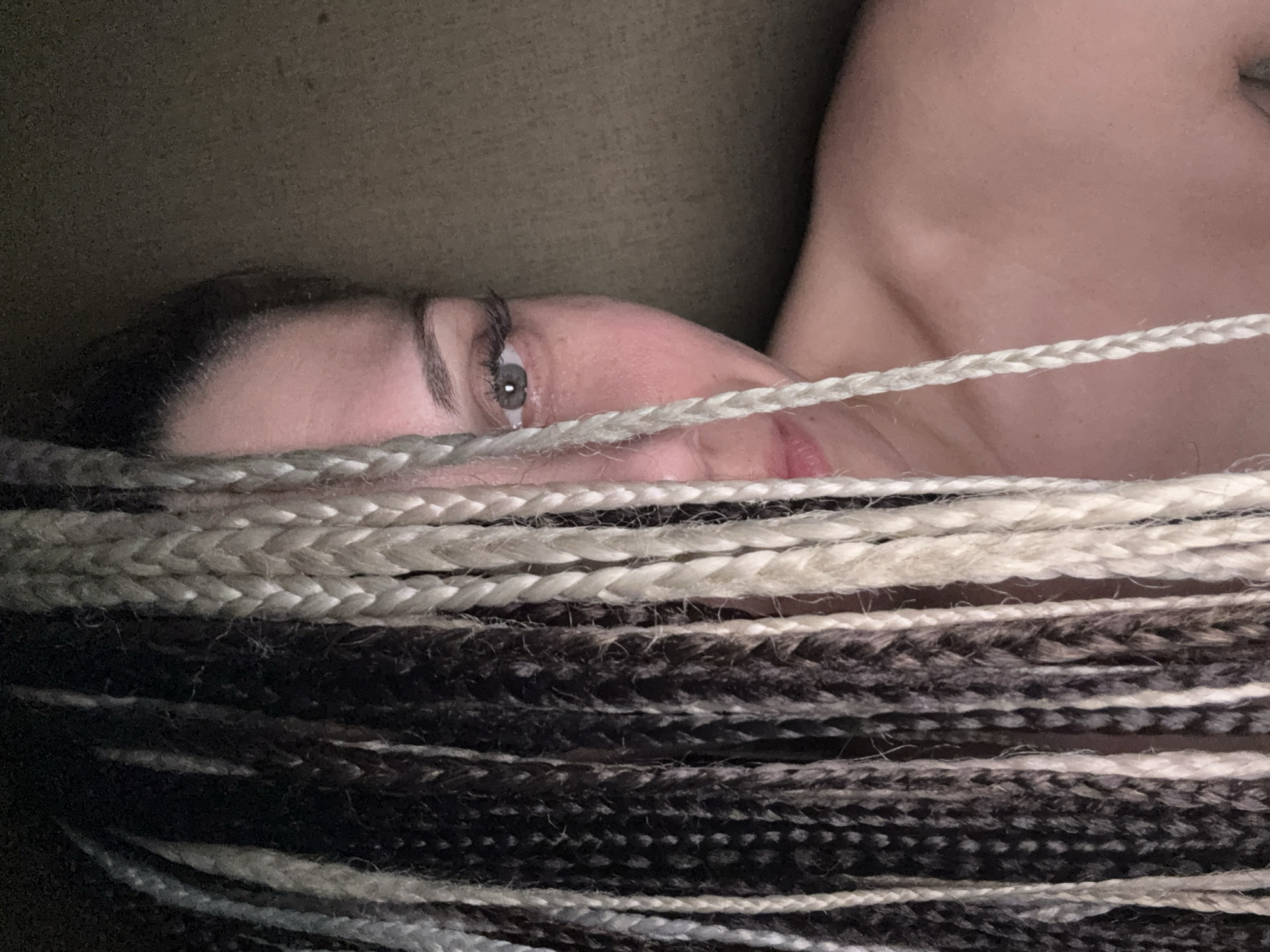 A close-up, horizontally oriented portrait features a person, likely female, resting indoors. The subject's face is partially visible in the middle-left, viewed from an upward angle, showing her right eye with a direct gaze, a well-defined eyebrow, a portion of her nose, and her upper lip. She wears long, multi-toned braided hair (possibly box braids or similar extensions) in shades of blonde/white, light brown, dark brown, and black. These braids create strong horizontal lines across the foreground, partially obscuring her face and upper body.
Her shoulder and upper chest are visible on the right side of the frame, suggesting she is lying down on a soft surface, possibly a sofa or bed. The background is a dark, indistinct texture, indicating an indoor setting with subdued lighting, perhaps evening or night. The person appears to be resting or posing for the photograph. There is no visible text in the image. The location context provided is Rusanovo, Russia, but no specific outdoor or architectural cues are visible within the frame.