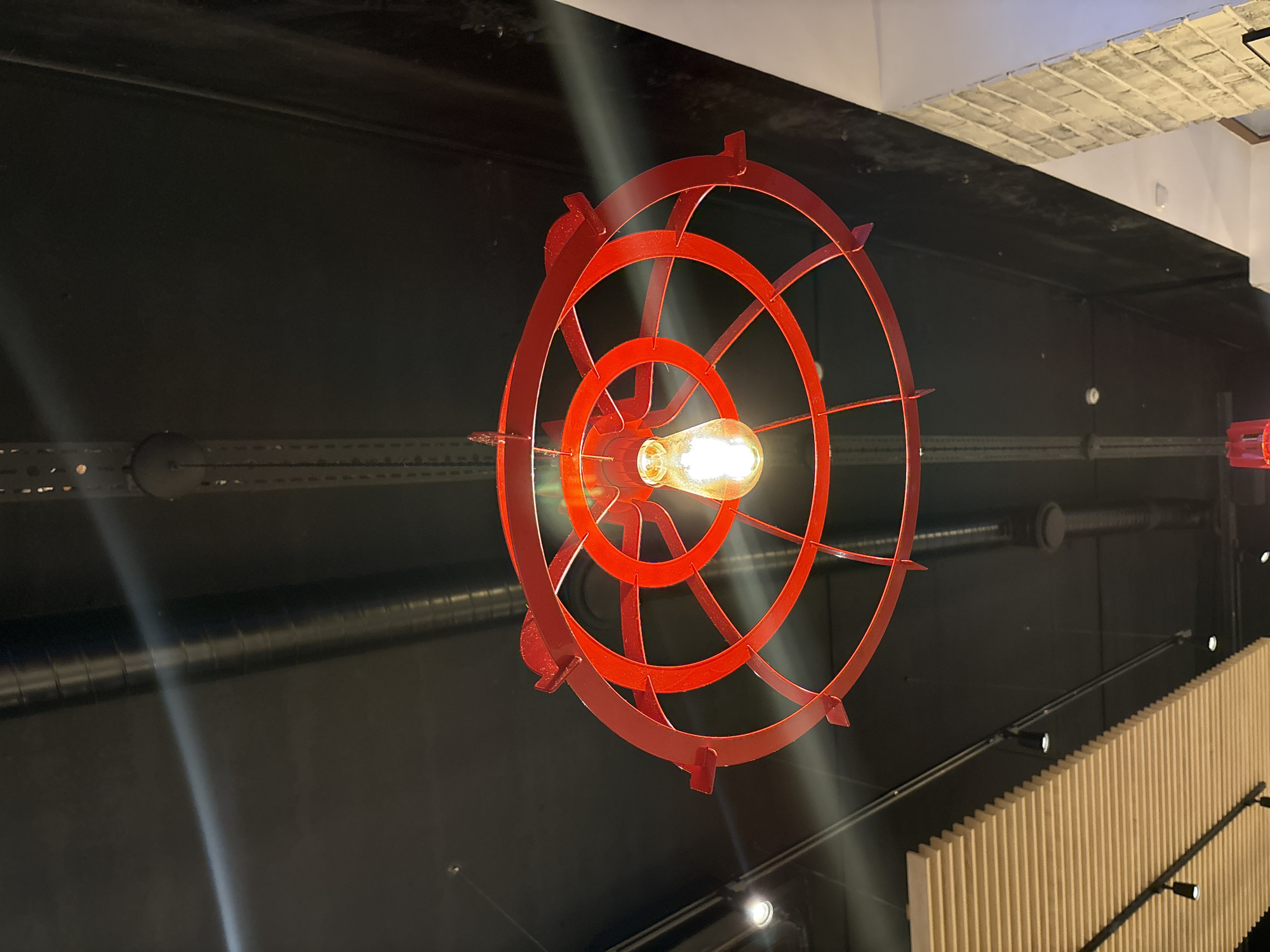 The image captures a striking interior scene, centered around a unique red light fixture. This fixture, designed in a circular, spoked pattern reminiscent of a ship's wheel or a stylized gear, hangs against a dark background. At its core, a glowing Edison-style light bulb illuminates the space, creating prominent lens flares and light streaks that cut across the frame.
The ceiling appears to be dark, possibly painted black, with exposed industrial elements including horizontal metallic pipes or ventilation ducts. In the upper right section, a patch of white or light grey square tiles with a distinct texture is visible on the ceiling. Towards the bottom right, a contemporary wall feature made of light-colored, vertically slatted wooden panels can be seen. Above these panels, a rail holds several small, bright spotlights, illuminating the textured surface.
The setting is indoors, suggesting a modern commercial space like a restaurant or bar, given the blend of industrial and warm design elements. The prominent artificial light source indicates that the photograph was taken in a dimly lit environment, likely during the evening or night, in Chaikovsky, Russia. There are no people visible, and no specific activity is taking place. No text is discernable in the image.