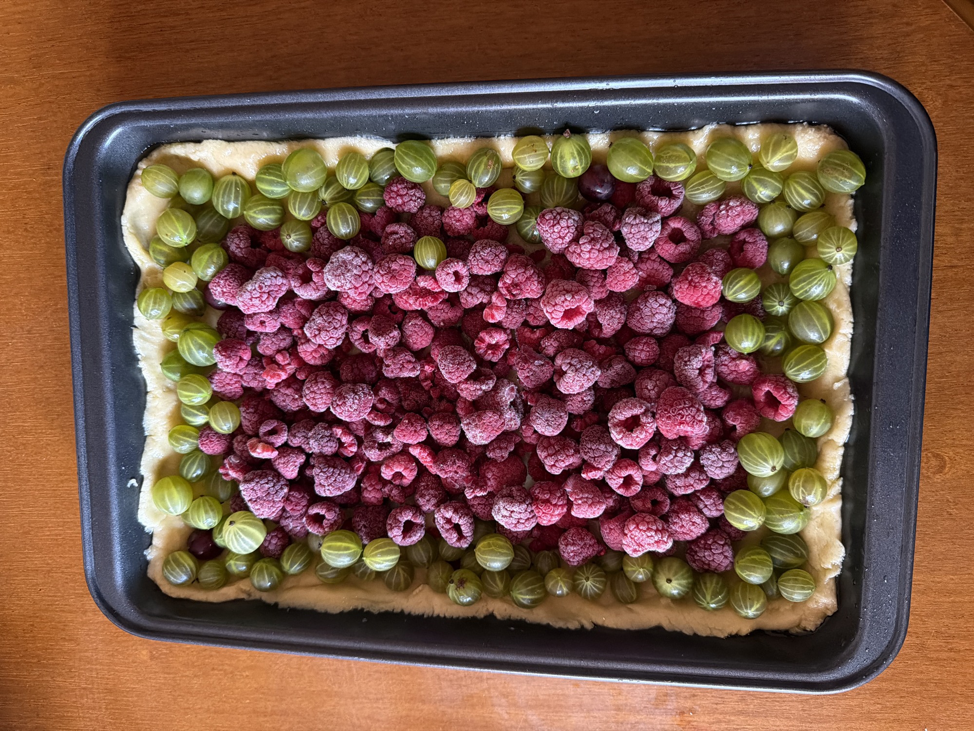 An overhead shot shows a rectangular black baking pan filled with raspberries and gooseberries on a bed of pastry. The berries are arranged in distinct sections, with a large pile of raspberries in the center, surrounded by a border of green gooseberries. The baking pan is placed on a wooden surface. There are no people or any context of Moscow, Russia present in the image.