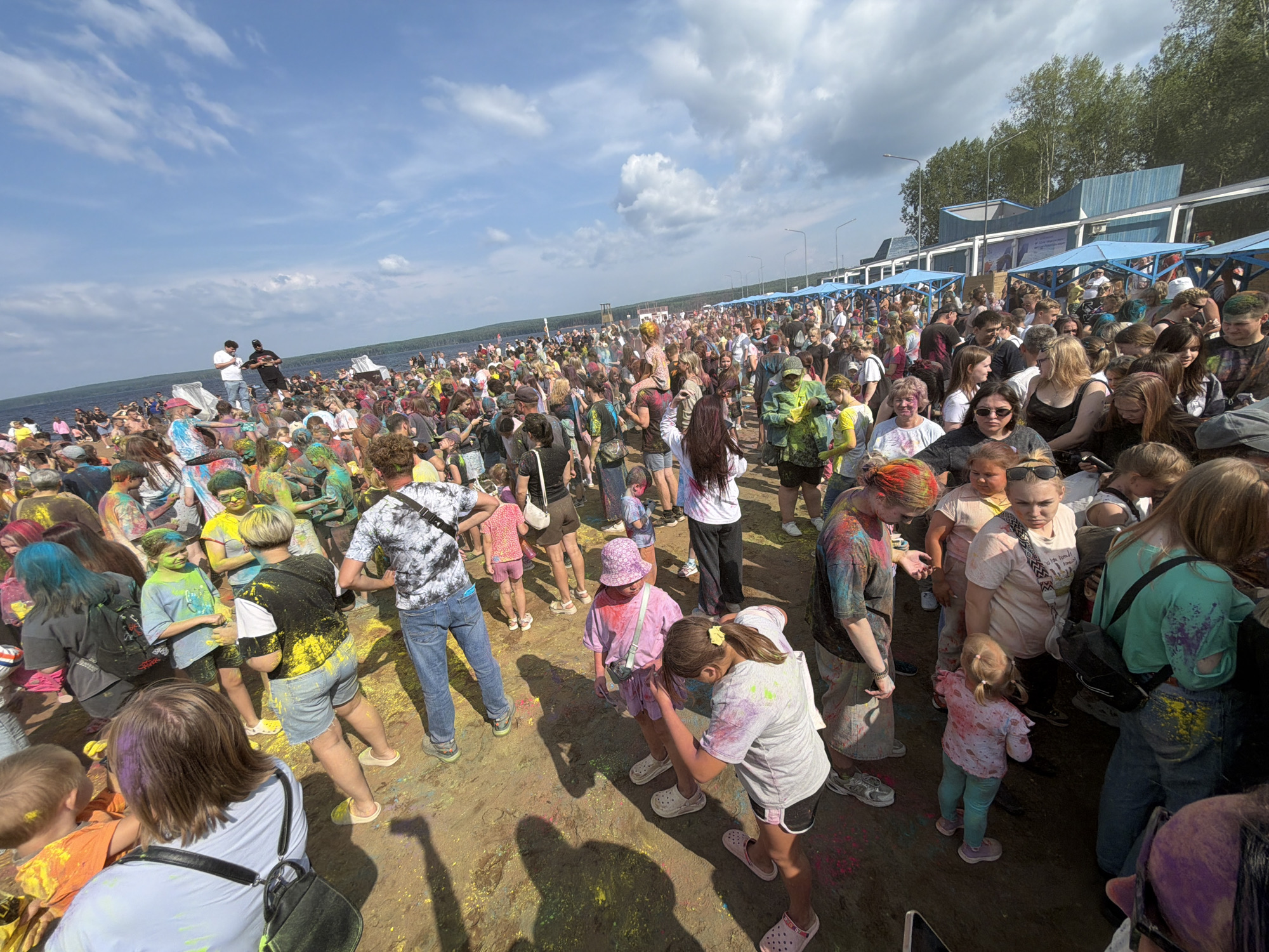 This image depicts a large outdoor festival, likely a color run or Holi celebration, taking place on a sandy area adjacent to a body of water, possibly the Kama River, in Chaikovsky, Russia. A dense crowd of people, diverse in age and attire, are gathered. Many are visibly covered in colorful powders, indicating participation in the event. The sky is partly cloudy, suggesting a bright, sunny day. In the background, structures resembling event booths or stalls with blue awnings are visible, along with trees lining the horizon. The overall atmosphere appears festive and energetic.