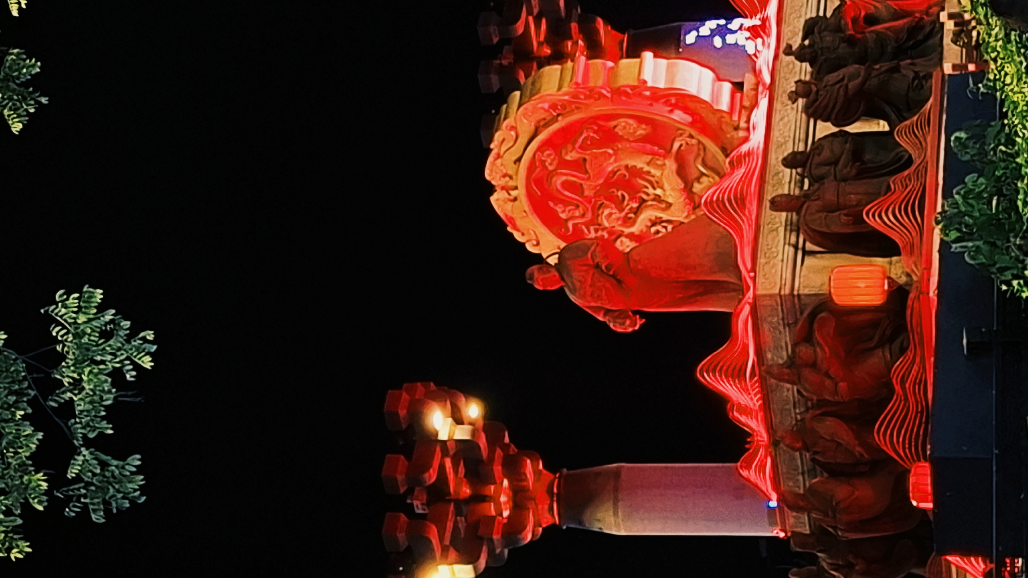 A night scene shows a portion of a brightly lit structure against a dark sky, possibly in Yanta District, China. The structure features elaborate details, including a large, circular red design with a golden dragon motif, suggesting a cultural or historical significance. Below this circular design are smaller, intricate red patterns and what appears to be a series of dark-colored figures, possibly statues, arranged along a vertical surface. The figures are punctuated with small, illuminated lanterns. A tree with green leaves is visible on the left side, further adding to the composition. The overall lighting is dramatic, with the red color dominating the scene and creating a visually striking contrast against the dark background.