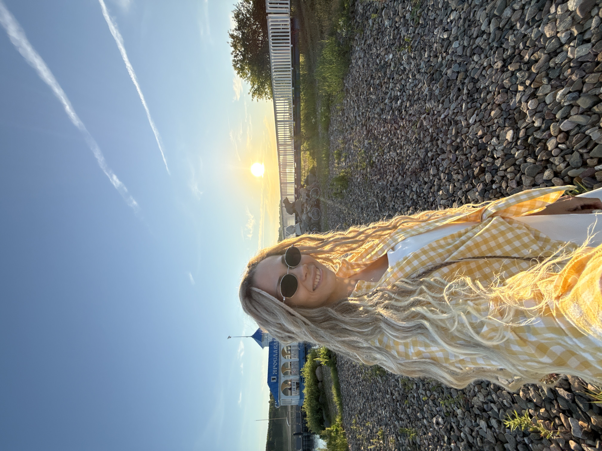 An outdoor scene features a woman with long, blonde dreadlocks and dark oval sunglasses smiling directly at the camera. She is wearing a white top beneath a yellow and white gingham-patterned shirt tied at the front. The setting sun, low on the horizon, backlights her hair and casts a warm, golden glow across the scene. The foreground and midground are covered in numerous small, grey-brown pebbles.
In the midground, to the left of the woman, a blue and white building with a decorative domed roof and multiple arches is situated next to a body of water. The Cyrillic word "ЯРОСЛАВЛЬ" (Yaroslavl) is prominently displayed in blue letters on the white facade of this building, confirming the location as Yaroslavl, Russia. To the right, a long, white, vertical-slatted fence or bridge extends horizontally across the scene. A dark, bear-shaped statue and two bicycles are visible near the fence, to the left of the sun. The sky is clear blue with two distinct white contrails from aircraft visible in the upper half of the frame. Green vegetation is scattered among the pebbles and around the base of the fence.