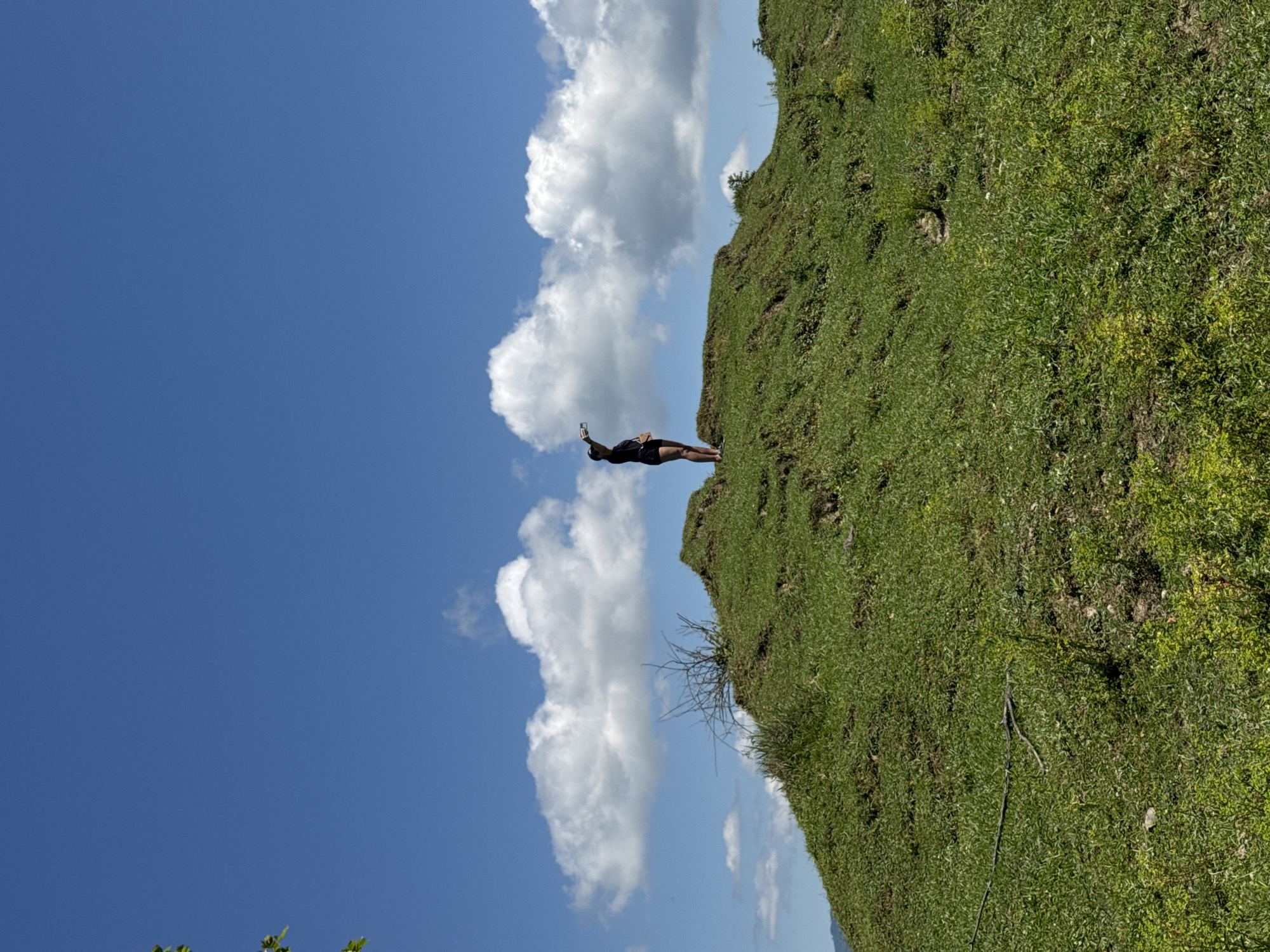 A person stands on a grassy knoll under a bright blue sky with scattered white clouds. The person, wearing a dark t-shirt and shorts, is facing away from the camera and appears to be holding something in their raised right hand. The knoll is covered in green grass with some sparse, dry vegetation. In the distance, a hillside can be seen with hints of foliage. The scene suggests an outdoor setting in a rural or natural area, possibly on the outskirts of Darcheli, Georgia.