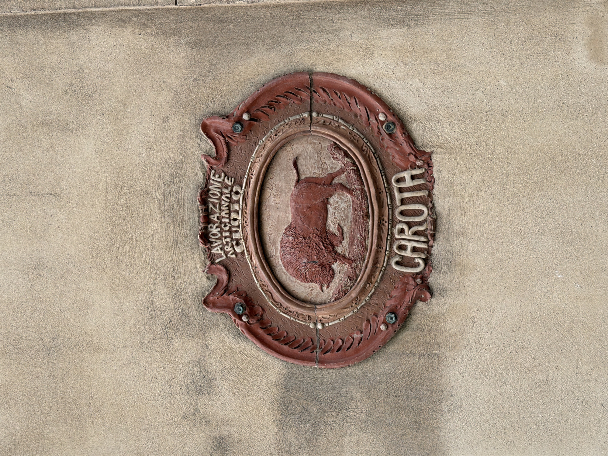 A close-up shot shows an ornate, oval-shaped, red-brown plaque affixed to a textured, beige wall. The plaque appears to be made of a cast material, possibly plaster or a similar substance. Within the oval frame is a depiction of a bovine animal, possibly a bull, in relief. To the left of the bovine figure is the vertical inscription "Lavorazione Artigianale", while to the right is the word "Carota" displayed vertically.
Given the linguistic context and visual elements, it is inferred that this plaque may be located in the city of Catania, Italy, as "Lavorazione Artigianale" is Italian for "Handcrafted". The plaque could be a sign for a business or a historical marker.