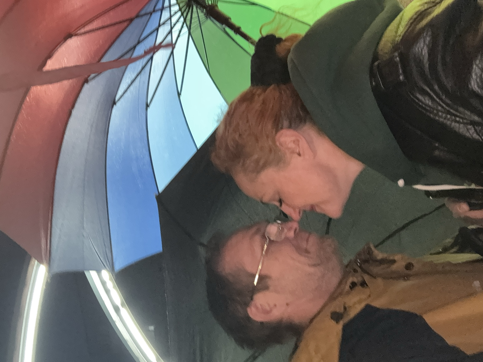 A couple is standing under a rainbow umbrella. The woman has red hair and is wearing a green hoodie. The man has glasses, and a beard, and is wearing a brown jacket. They appear to be about to kiss. The scene appears to be at night or in low light, based on the ambient lighting and the presence of lights in the background. The location is not discernible, as there is no visible reference to Chaikovsky, Russia.