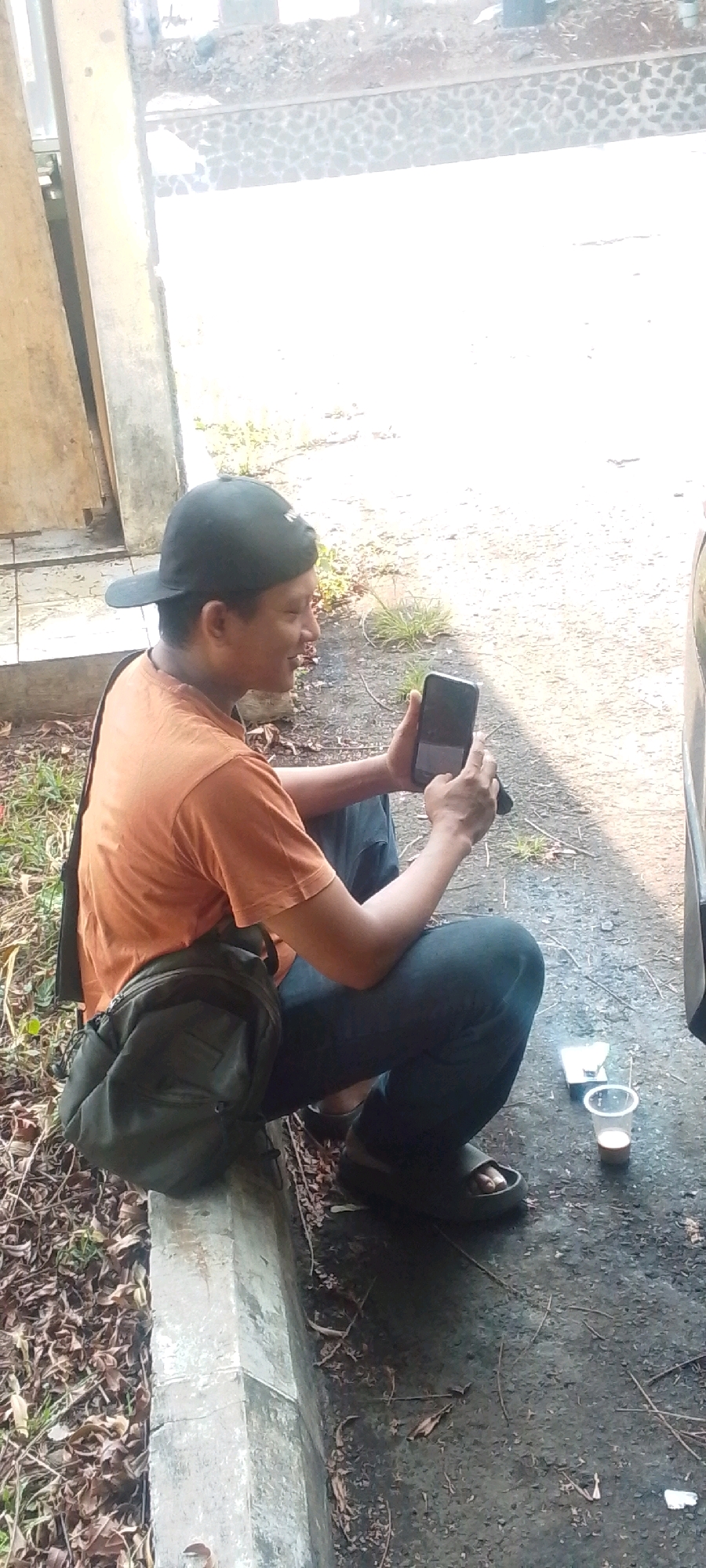 A man in an orange t-shirt and blue jeans is crouching outdoors on a cracked concrete curb in Cilaku, Indonesia. He wears a black baseball cap and dark sandals. A dark green messenger bag is slung across his left shoulder. In his hands, he holds a black smartphone, which he is looking at. To his right, on the ground, is a small white object and a clear plastic cup containing a light brown liquid. The ground is composed of asphalt and dirt, littered with dry leaves and twigs. Behind him, a rough stone wall borders an area of dry earth and scattered debris. The scene is bathed in bright sunlight, casting distinct shadows.