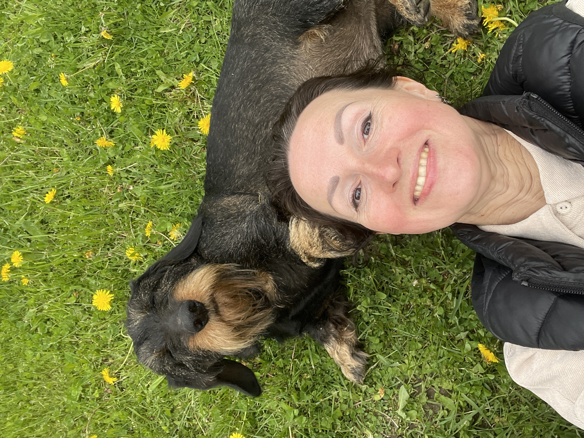 A woman lies on her back on grass with yellow dandelions. A dachshund dog lies beside her. The woman is smiling. She is wearing a white shirt and black puffer jacket. The dog has dark brown and tan fur. The location is unknown.