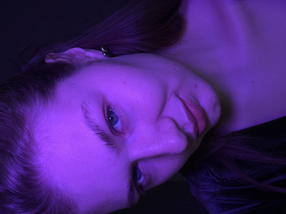 A person's face is illuminated with a violet light, angled slightly to the side. The subject has fair skin, blue eyes, and light reddish-brown hair. An earring is visible in their left ear. The mouth is slightly open, revealing pink lips. The person's head is tilted on a dark surface, possibly fabric. The lighting creates deep shadows, particularly around the hair and neck area.