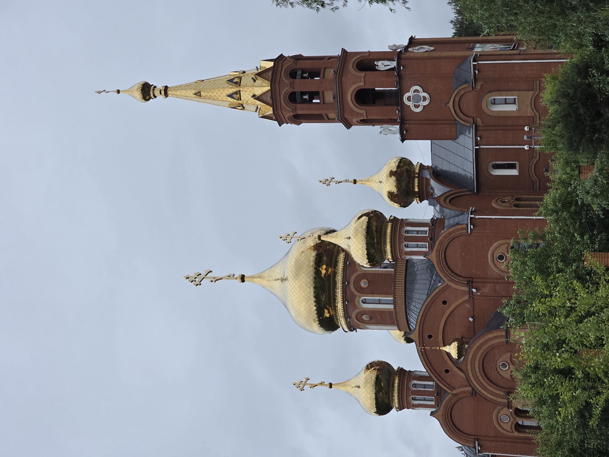 This image depicts the St. George the Victorious Cathedral, located in Chaikovsky, Perm Krai, Russia, under an overcast sky. The cathedral is constructed predominantly from red brick, featuring a complex architectural design. Five large, golden onion domes are prominent, with each capped by a gilded Orthodox cross. A smaller, golden onion dome is situated centrally on the lower frontal section, also surmounted by a cross. To the right, a tall, slender bell tower rises, culminating in a golden, intricately patterned spire topped with an Orthodox cross. The bell tower includes multiple arched belfry openings where bells are visible, and its brickwork is accented with a white quatrefoil emblem and two white statuettes, resembling angels, positioned on ledges below the belfry. The lower portions of the cathedral are partially obscured by dense green deciduous trees and bushes in the immediate foreground. The sky is uniformly light grey, indicating an overcast condition. No human figures, animals, or overt actions are present in the scene.
