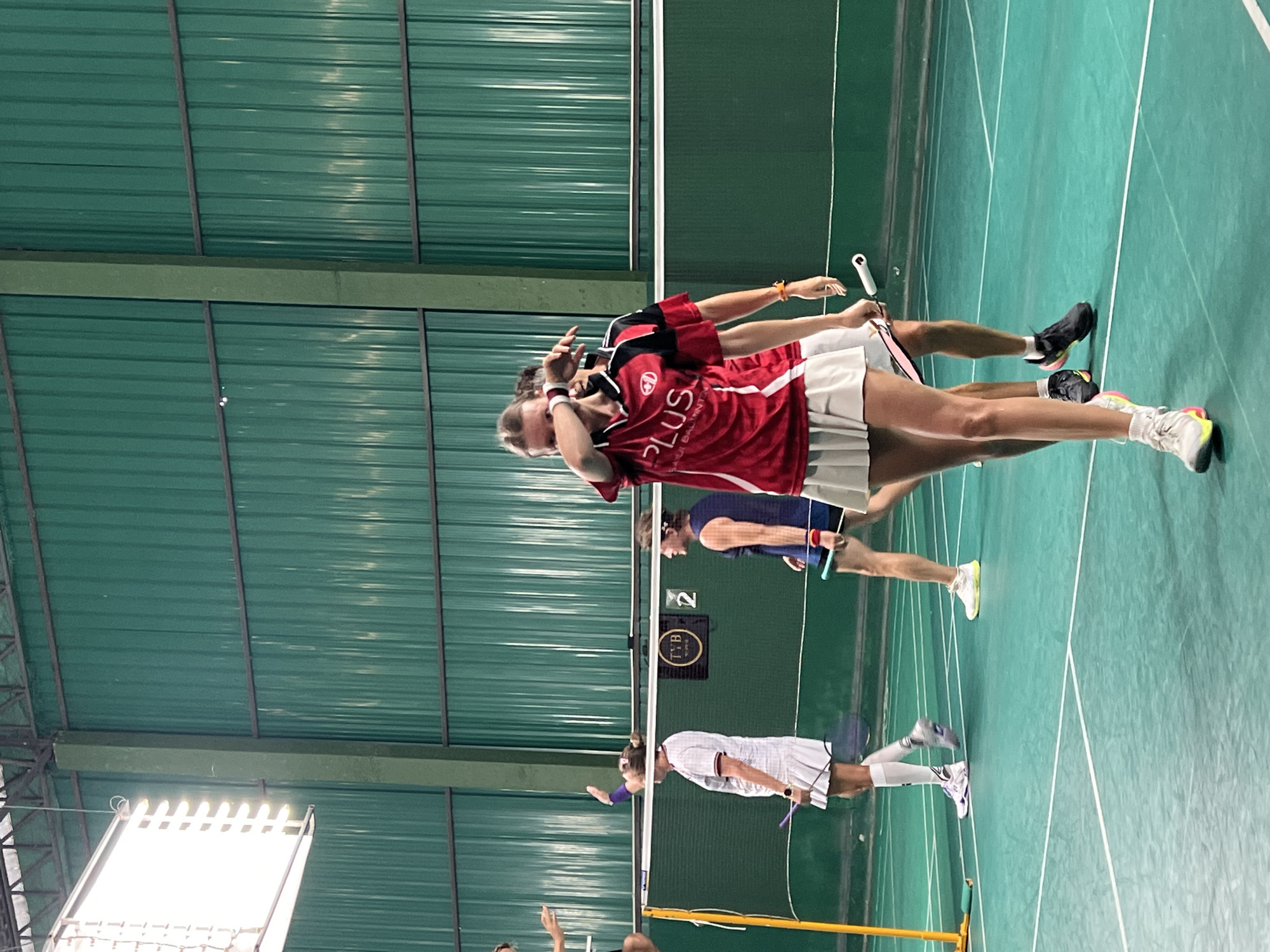 The image depicts an indoor badminton court in Ban Bang Rak, Thailand. The court surface is green with white lines. In the foreground, a female badminton player in a red and black jersey and a white pleated skirt is visible. She is holding her hand up to her face, possibly wiping sweat or shielding her eyes. Another player in a blue top and white skirt is in the middle ground, facing away from the camera. Behind them, a net spans the court. The background features green corrugated walls and bright fluorescent lighting. Several other players and badminton rackets are also present on the court.