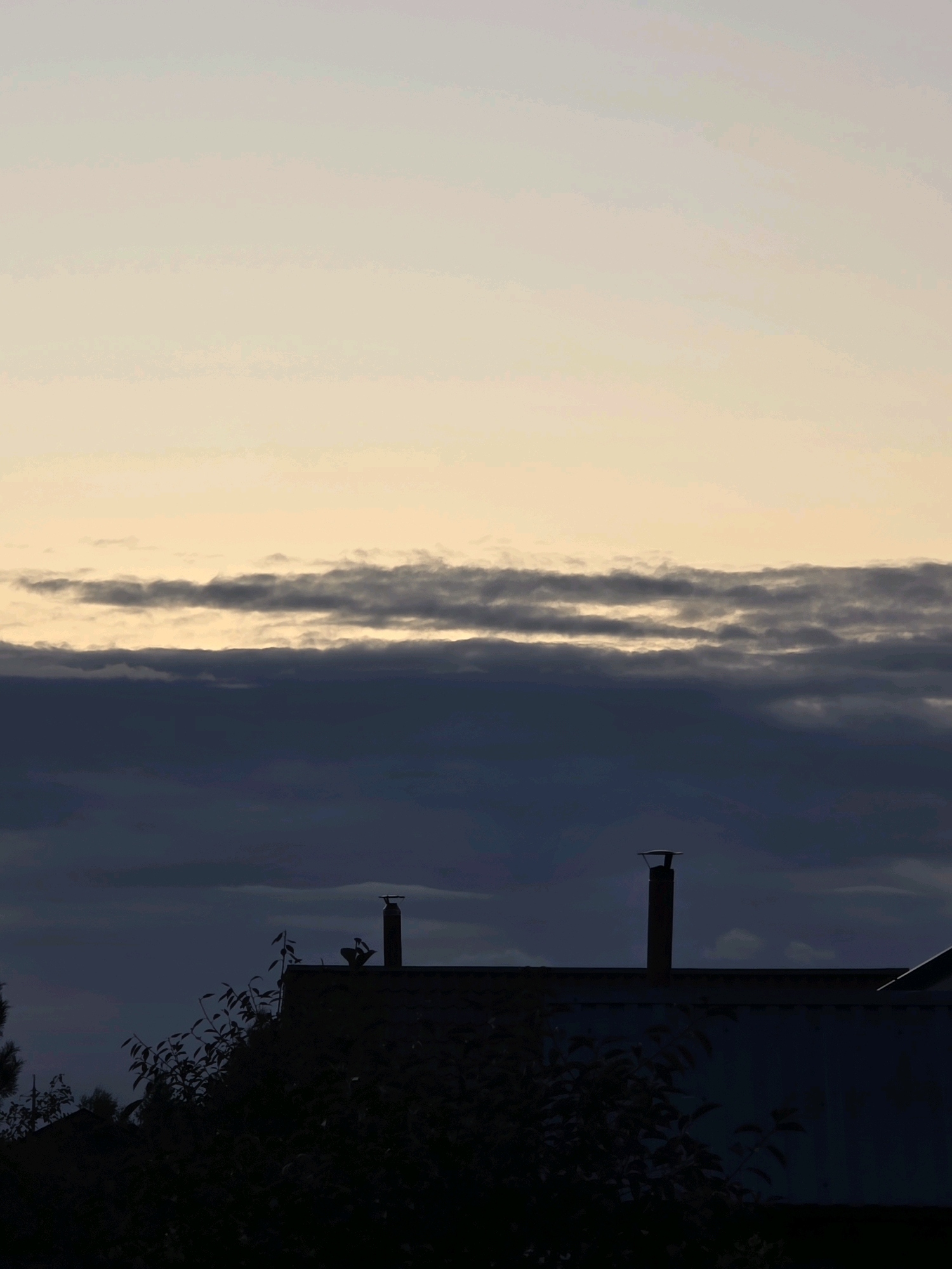 The image shows a silhouetted view of rooftops against a twilight sky. The sky transitions from a pale yellow at the horizon to a light blue at the top, with horizontal bands of dark clouds obscuring the view. Two chimney stacks are visible on the roof of a building in the foreground, with one appearing taller than the other. Silhouetted foliage from trees partially covers the lower portion of the image, suggesting a residential area. The scene appears to be captured in the city of Речицы, Russia, during dusk.