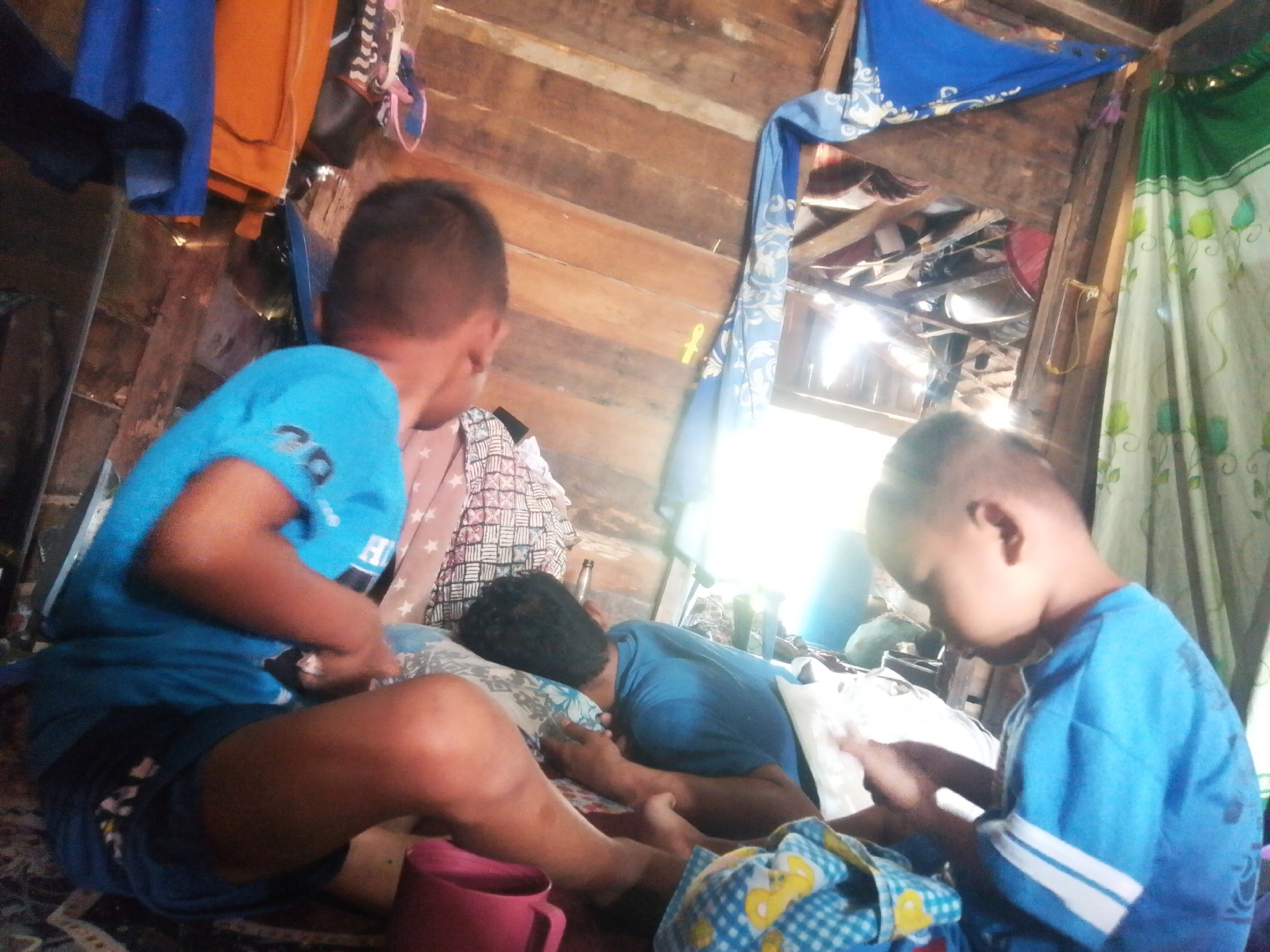 In a dimly lit, rustic interior, likely a dwelling in Labuhan Bilik, Indonesia, three young boys are partially visible. Two boys, wearing blue t-shirts, are the primary subjects. The boy on the left is in the foreground, his back to the camera, with his head turned away. He appears to be sitting or lying down, wearing a blue t-shirt with white stripes on the sleeves. His legs are bent, and he is wearing dark shorts. Beside him, a patterned blanket with stars is visible. To his right, another boy, also in a blue t-shirt, is lying on his side facing away from the camera, with his head resting on a pillow. He is wearing a blue t-shirt. A third boy, his face and torso visible, is also wearing a blue t-shirt with white stripes on the sleeves. He is looking down at something in his hands, possibly a phone or a small object.

The environment suggests a simple, possibly unfinished structure with wooden walls and beams. Various fabrics are draped and hung, including blue and orange textiles at the top left, a blue patterned cloth hanging from the ceiling on the right, and a green and white floral curtain on the far right. Sunlight streams in from an unseen source, creating bright highlights and deep shadows. A red mug is visible near the foreground boy's legs. The overall impression is a glimpse into a casual, everyday scene within a humble home.
