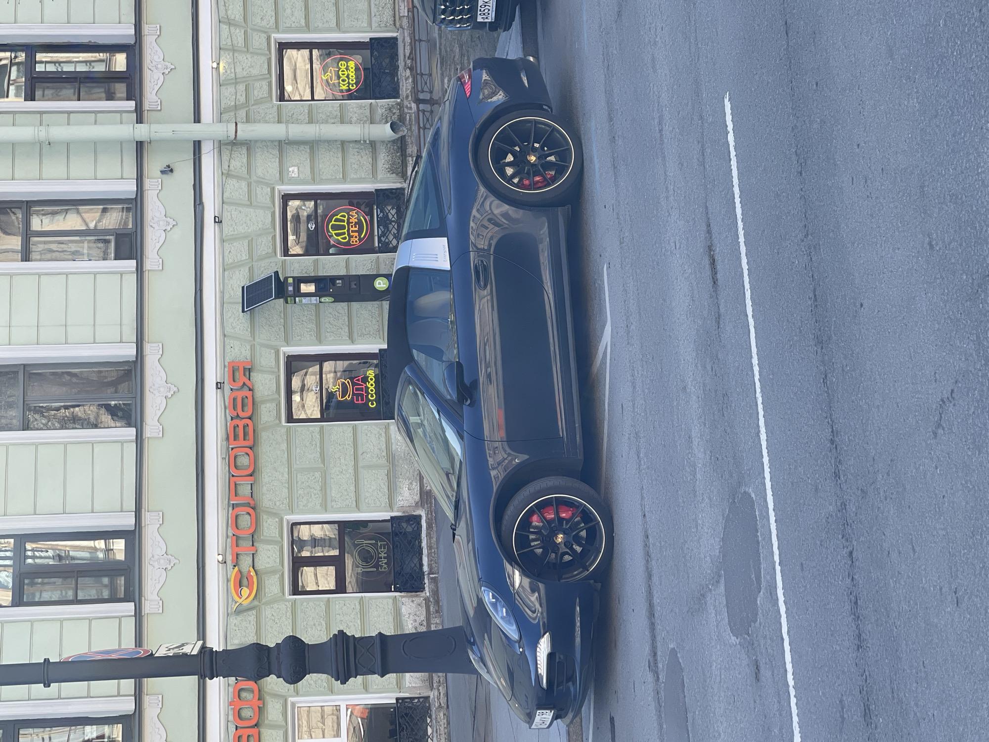 An upright, eye-level shot captures a black Porsche 911 Targa parked on an asphalt street in an urban setting. The sports car, featuring black multi-spoke wheels with a polished rim and prominent red brake calipers, is positioned parallel to a multi-story building. Its distinct silver Targa roll bar and "Targa" badge are visible on the side.
The building behind the car has a light green or grey facade, adorned with white, ornate architectural details around its windows. Several windows display neon signs, primarily in Russian, advertising "КОФЕ с собой" (COFFEE to go), "ВЫПЕЧКА" (BAKERY), "ЕДА с собой" (FOOD to go), and "БАНКЕТ" (BANQUET). A large, red horizontal sign on the building, partially obscured by a black lamppost, reads "СТОЛОВАЯ" (CANTEEN/DINING ROOM). A parking meter or device with a solar panel is affixed to a pole near the car.
The street features a single white dashed line marking. The overall impression is that of a bright, clear daytime, suggesting good weather. The scene depicts a typical street view in Saint Petersburg, Russia, given the visible Russian text and architectural style.