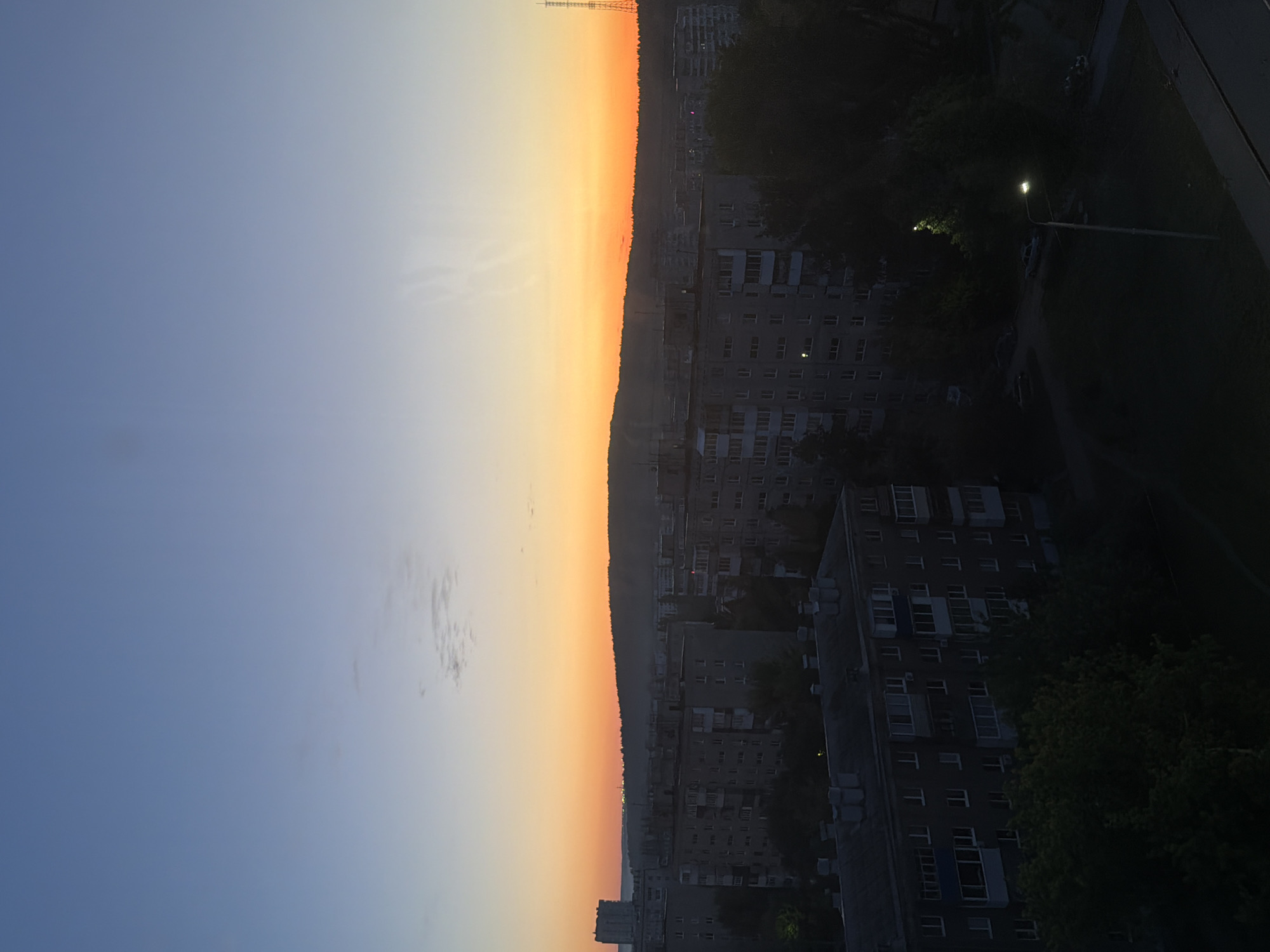 The image shows a city skyline at sunset. The sky transitions from blue to orange, with the sun setting over the horizon. In the foreground, there are several buildings, mostly apartment complexes, silhouetted against the sky. Streetlights are visible in the dark, treed areas between buildings. A radio tower can be seen near the horizon. The location is Unknown, Unknown.