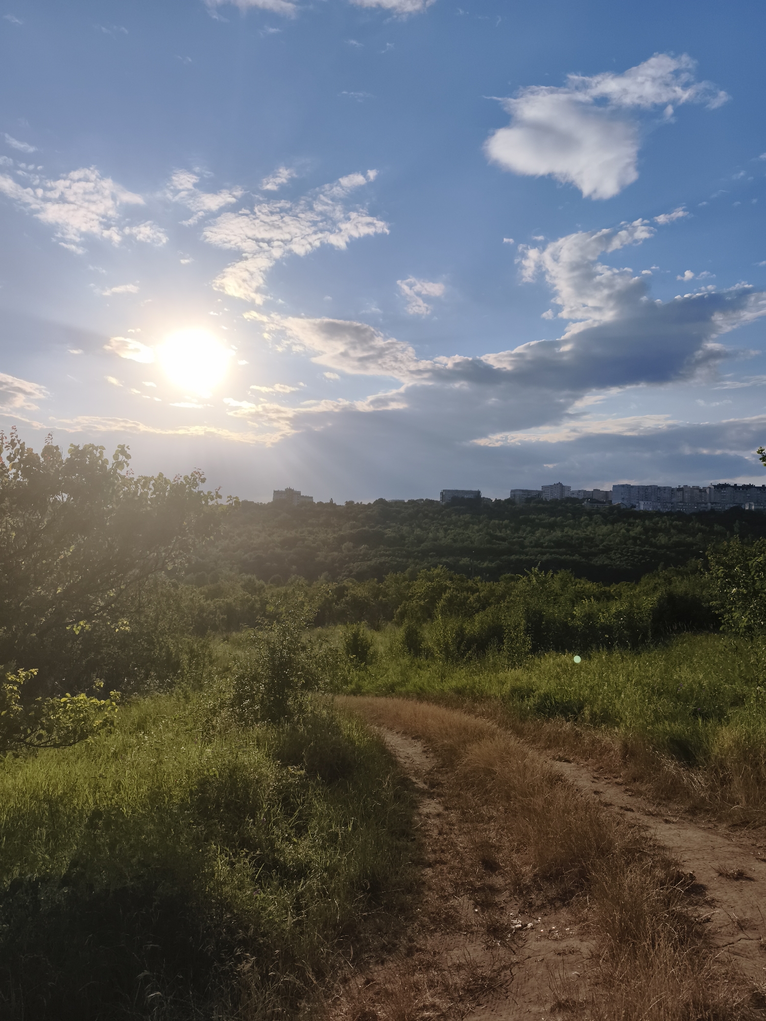 The image depicts a rural landscape with a dirt road in the foreground, curving to the right. The road is flanked by tall green grass and dry, brown vegetation. In the mid-ground, a dense forest covers rolling hills. Beyond the forest, a cityscape with numerous apartment buildings is visible on the horizon. The sky is a vibrant blue with scattered white and gray clouds. The sun is shining brightly, casting rays of light through the clouds and illuminating the scene. The overall impression is one of natural beauty transitioning into an urban environment. The location is identified as Codru, Moldova. There are no people or animals visible in the image.