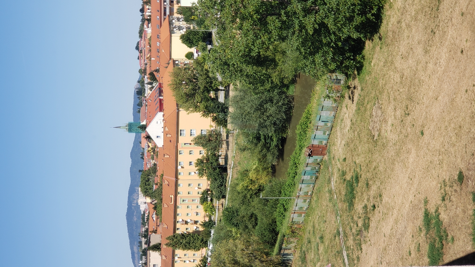 The image depicts a cityscape in Prešov, Slovakia, on a clear, sunny day. In the foreground, a patch of dry, yellowed grass is visible, with a weathered green fence running horizontally across the middle ground. Beyond the fence, a narrow, slow-moving body of water, likely a river or canal, is present, lined with dense green foliage.
In the middle ground, a row of apartment buildings with orange-tiled roofs and light yellow facades stretches across the scene. These buildings feature numerous uniformly spaced windows and balconies. Scattered trees, some evergreen and some deciduous, are interspersed between and in front of the buildings. A single lamppost stands near the fence.
The background reveals more urban development with buildings of varying heights and architectural styles, predominantly featuring orange or red tiled roofs. A prominent green spire, possibly belonging to a church or other public building, rises above the rooftops, and appears to be partially covered in scaffolding or netting. In the far distance, rolling hills or mountains are silhouetted against the bright blue sky. No people are visible in the image. The overall impression is one of a residential area within a city, with a natural element (the waterway and vegetation) integrated into the urban landscape.