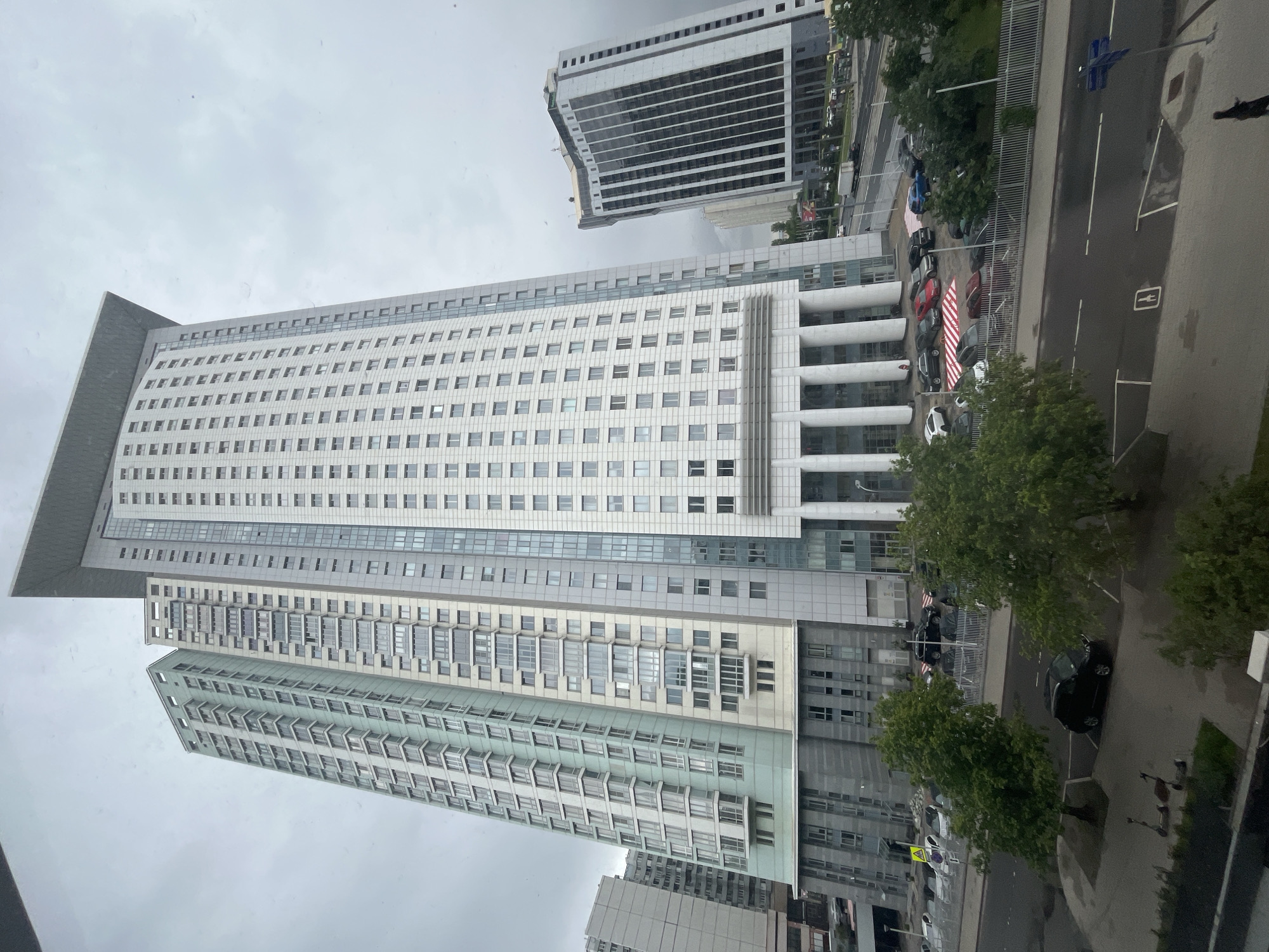 The image shows a view of Moscow, Russia, likely taken from a high vantage point such as a window in a building. The main subject is a tall, multi-story building with a large number of windows, a flat roof, and a rectangular shape. The building's facade is primarily white with gray accents. A second high-rise building is seen in the upper part of the frame. Adjacent to the main building is a parking lot with cars and trees. The sky is overcast. The image provides an urban setting focused on architecture and city infrastructure.