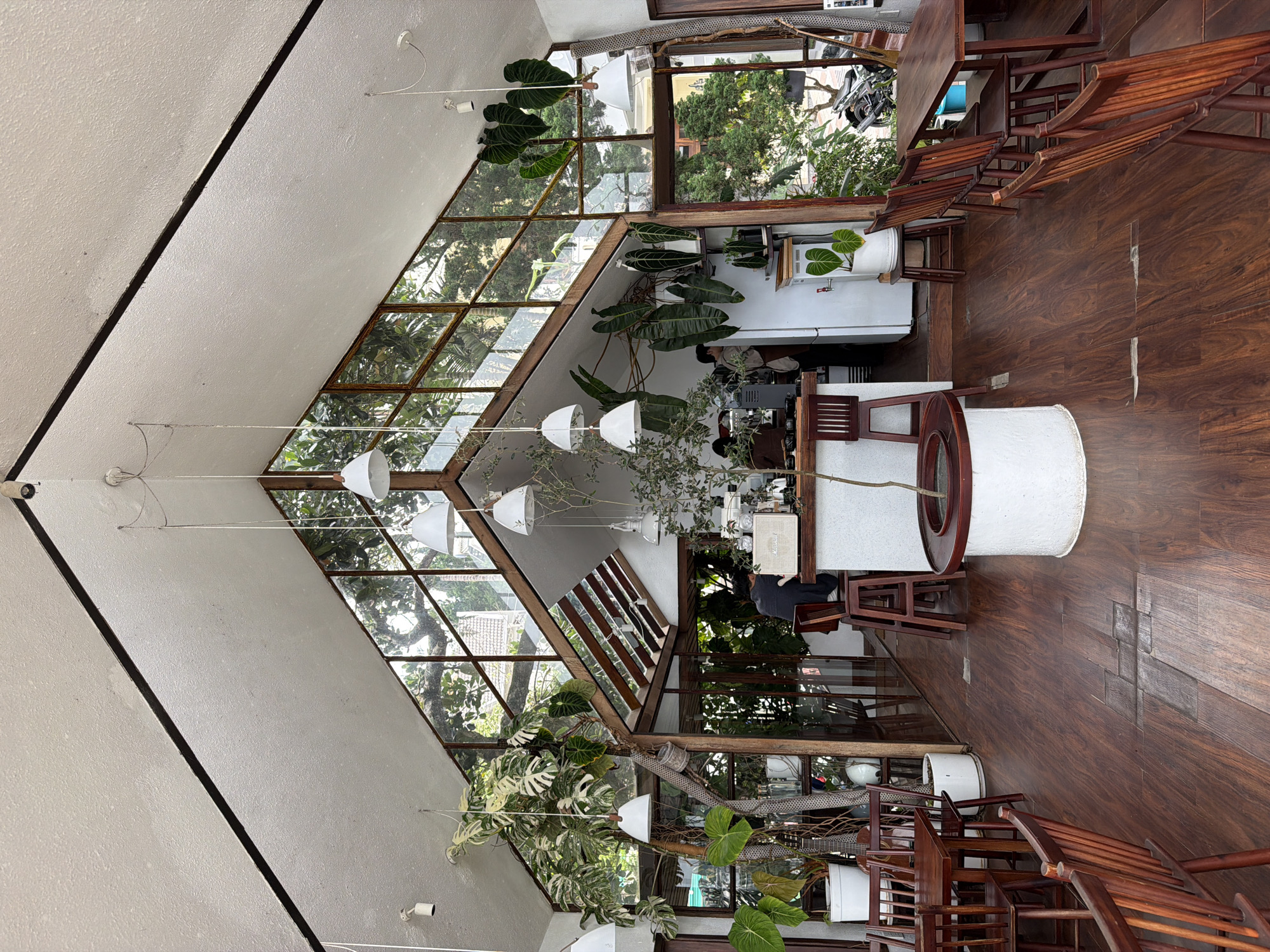 The image depicts the interior of a spacious, modern coffee shop in Da Lat, Vietnam, characterized by its high, angled ceiling with exposed wooden beams and expansive glass walls that allow ample natural light and offer views of the surrounding greenery. The flooring is dark, polished wood, adding to the warm ambiance.
The space is sparsely furnished with several wooden tables and chairs. In the foreground, to the left, a small cluster of tables and chairs are visible. Further into the shop, a counter area is set up, with a barista attending to tasks. A large Monstera plant, with its distinctive split leaves, is a prominent feature, adding a tropical element to the decor. Hanging pendant lights with white shades are strategically placed throughout the interior.
Various potted plants are scattered throughout the cafe, reinforcing the connection with nature. The overall atmosphere is serene and inviting, with a minimalist yet lush aesthetic. The clientele appears to be minimal, with a few individuals visible in the background, suggesting a quiet moment within the establishment.
