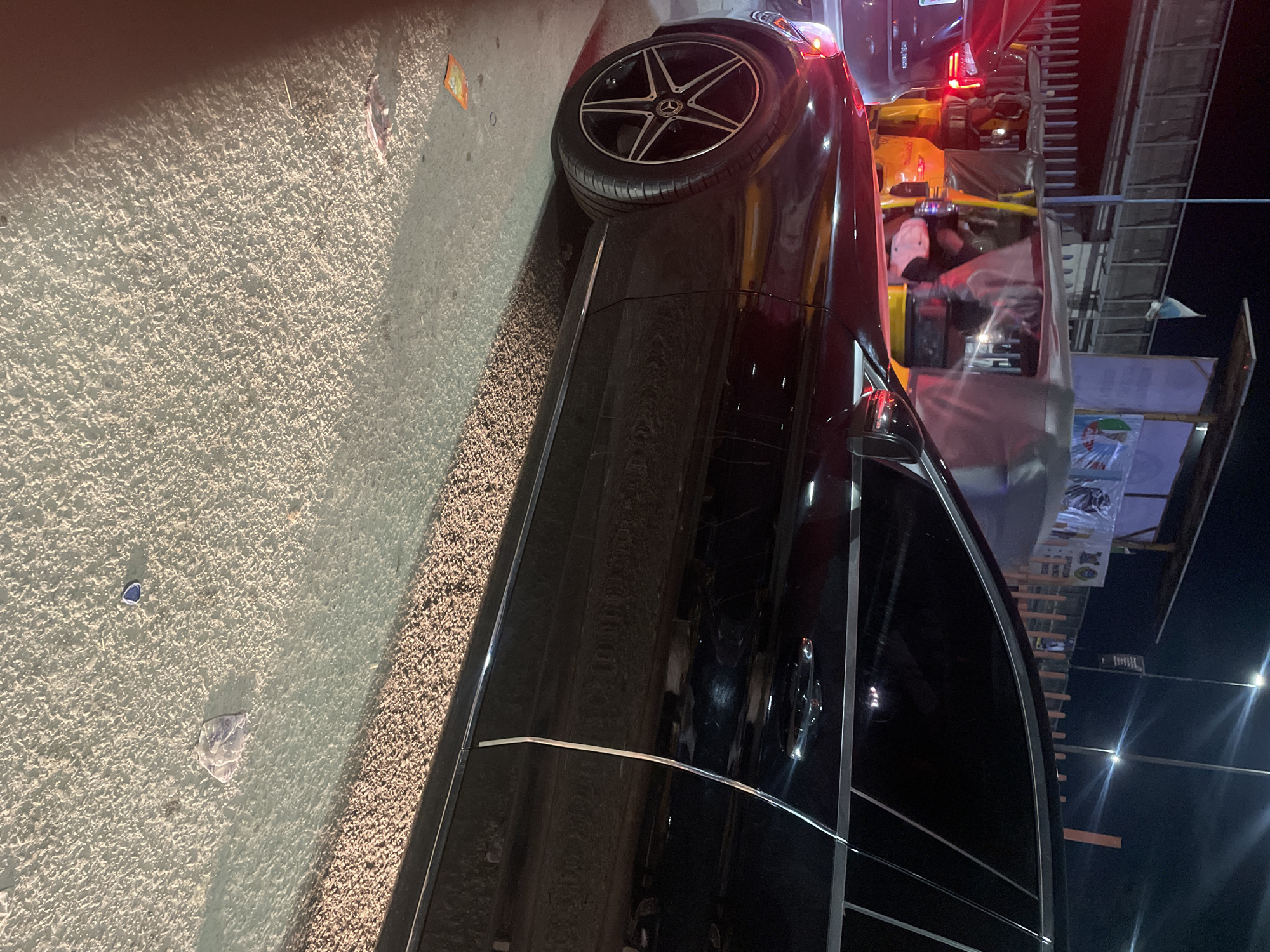 A black Mercedes-Benz is parked along a street in Lekki, Nigeria. Adjacent to the vehicle are commercial stalls, visible in the background. The street is illuminated by streetlights. Additional vehicles are nearby.
