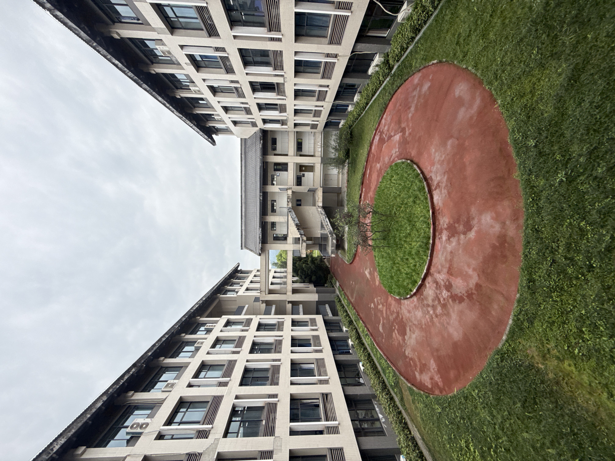 An upward-angled photograph captures the courtyard of an apartment complex. The courtyard contains a circular, red running track encircling a grassy area. Buildings line both sides of the courtyard. The sky is overcast. Based on the architectural style, the location is likely Jiangjin, China. No people are visible.