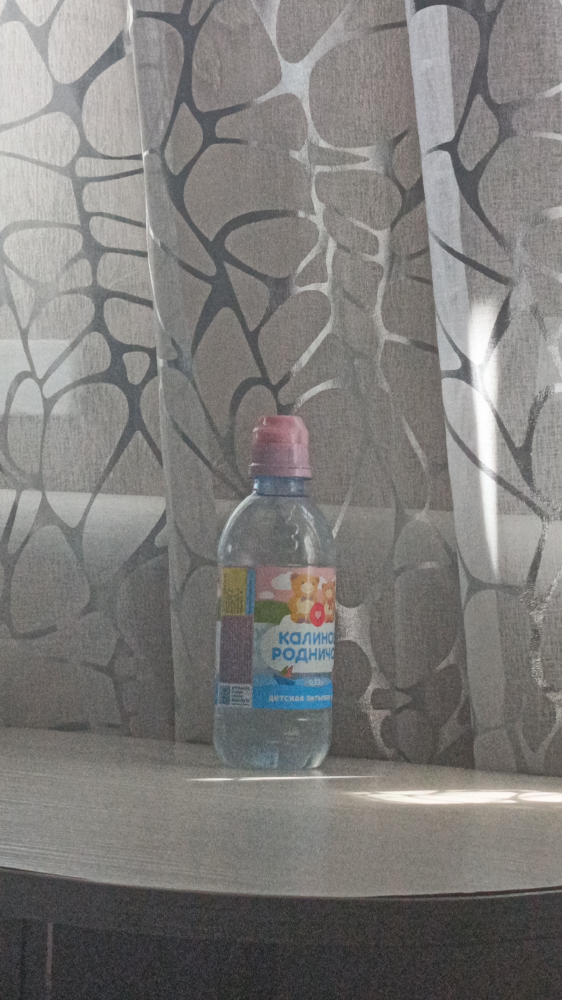 A clear plastic bottle of "Калино Родничо" (Kalino Rodnicho) children's drinking water, with a pink cap, stands on a surface in front of a grey patterned curtain. The label on the bottle features cartoon bears and text in Russian. The lighting suggests it is daytime, with sunlight casting a bright reflection on the surface. The overall scene is indoors, likely a domestic setting, in the village of Stanovoe, Russia.