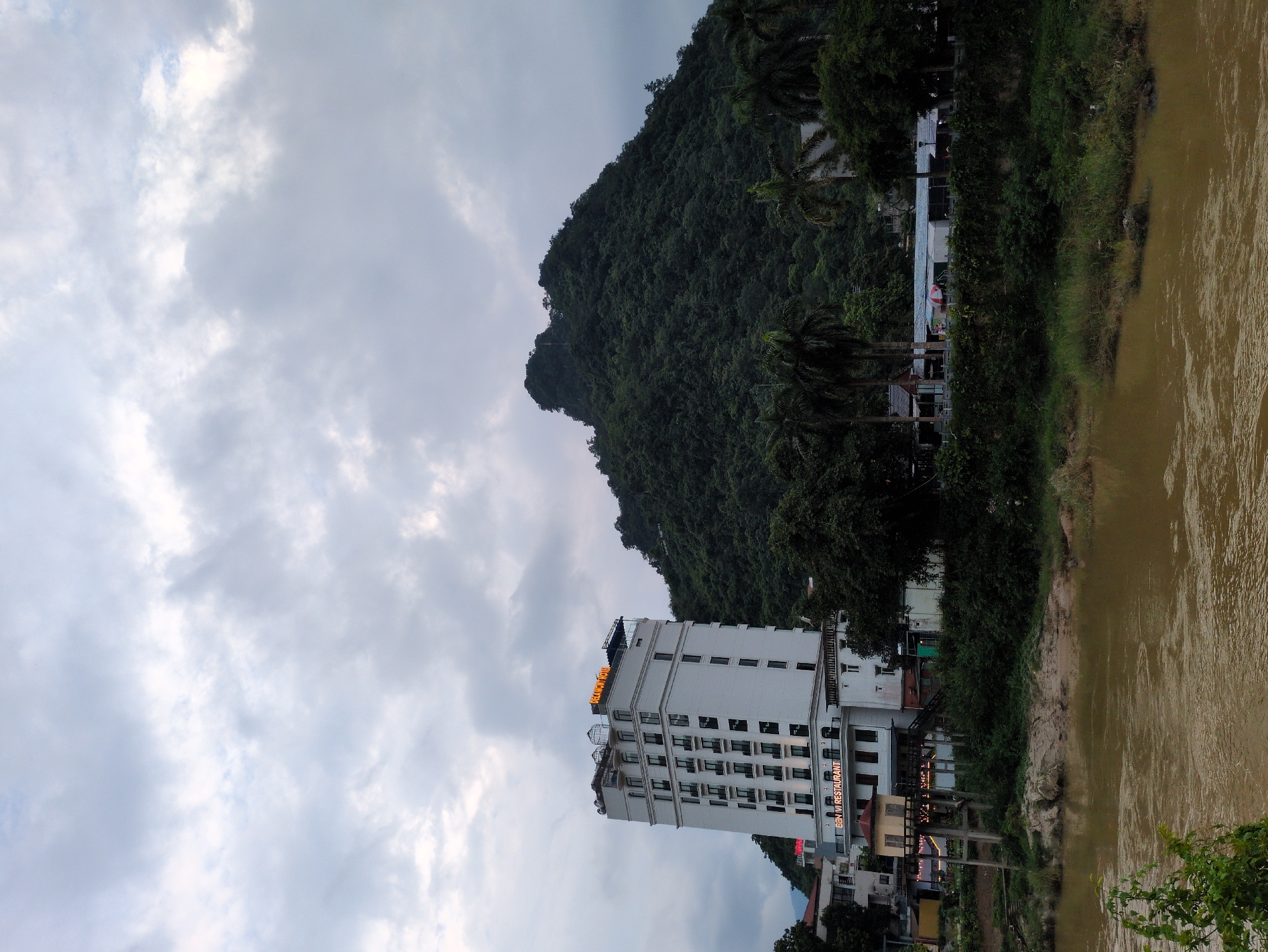 This image, captured in Ward 1, Hà Giang City, Vietnam, depicts a waterfront scene dominated by a multi-story white hotel, "DELAMONT HOTEL," and a lush, tree-covered mountain. The hotel, situated to the left, is a prominent structure with numerous windows and balconies, featuring illuminated signage. In front of the hotel, a restaurant named "BAM VI RESTAURANT" is visible. The mountain, a dark green mass of foliage, rises steeply behind the hotel and extends across the right side of the frame. In the foreground, a murky brown river flows, with a sandy bank visible on the left. Palm trees and other dense vegetation line the riverbank and the base of the mountain, creating a natural boundary between the built environment and the wild landscape. The sky is overcast with heavy, grey clouds, suggesting a muted or perhaps approaching rainy weather. There are no people clearly visible in the image. The overall scene conveys a sense of a developing urban area nestled against a dramatic natural backdrop.