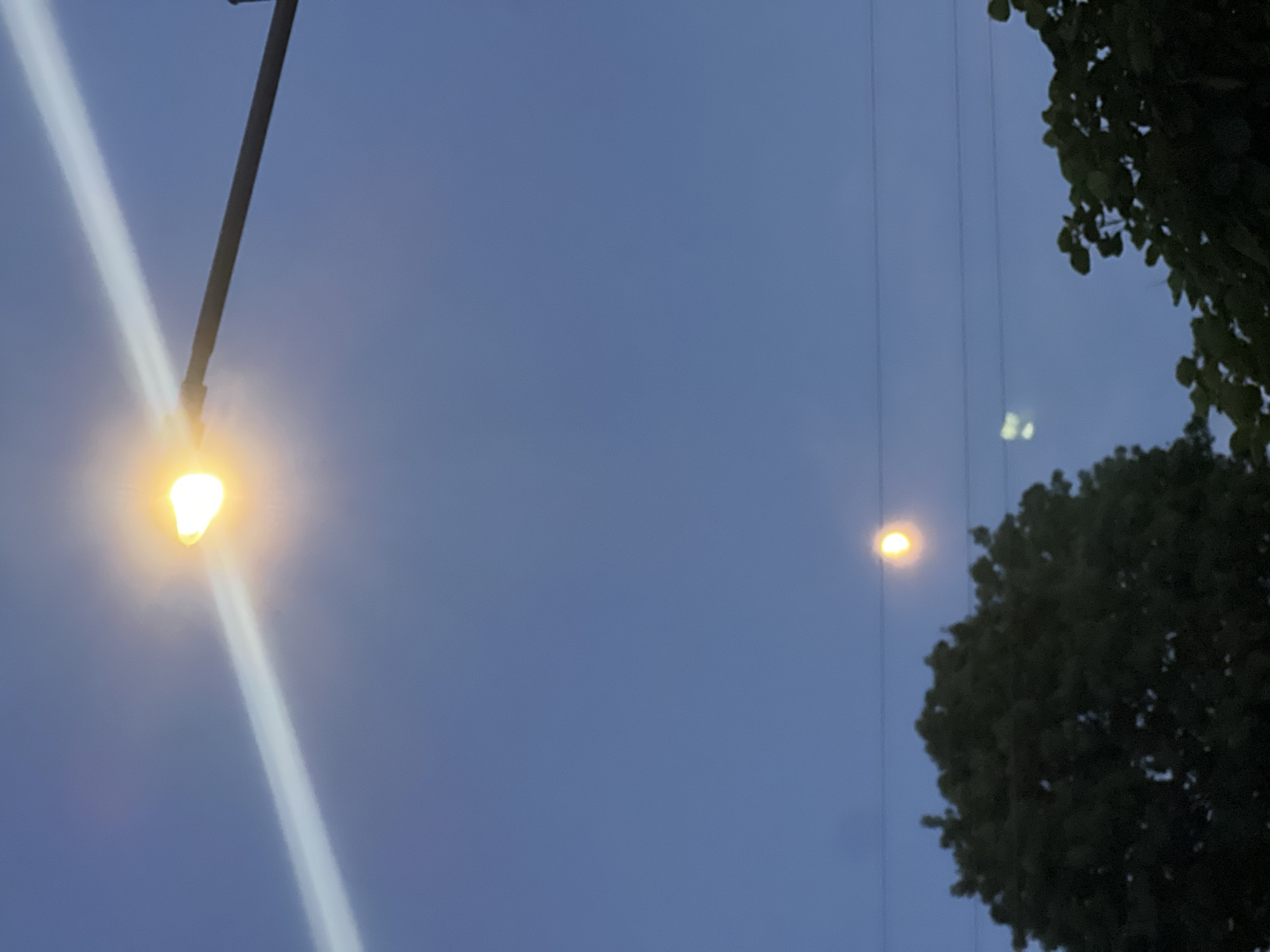 The image shows a twilight sky with a bright street lamp in the upper left quadrant, casting a visible beam of light diagonally across the frame. The lamp is attached to a dark pole. Further down, a second, dimmer light source is visible, possibly another street lamp or a reflection. Two indistinct dark shapes in the lower part of the image appear to be trees, one on the left and one on the right. Thin, dark horizontal lines, likely power lines, are visible across the sky. The sky is a deep blue, indicating it is either early morning or late evening. The overall scene depicts an outdoor environment in Kryzhanivka, Ukraine, likely a street or park area during dusk or dawn. No people are visible in the image.