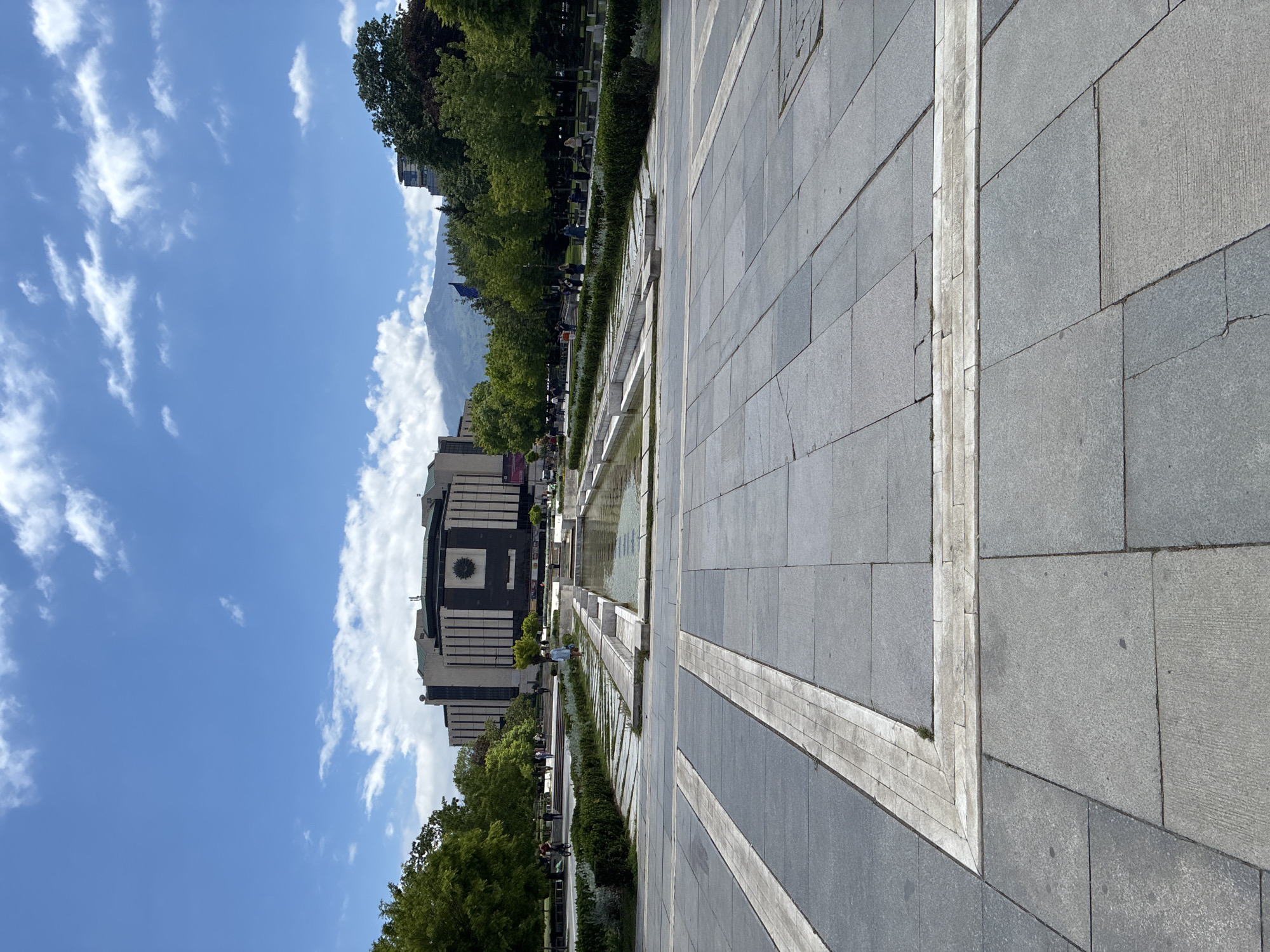 The image shows a cityscape in Sofia, Bulgaria. The National Palace of Culture stands prominently against a blue sky dotted with cumulus clouds. A mountain range is visible in the background, partially obscured by trees. In the foreground, a paved plaza made of gray stone tiles stretches out, featuring a linear water fountain with greenery along its edges. A few people are walking in the plaza, near the trees and fountain. The overall impression is of a sunny day in a modern urban space.