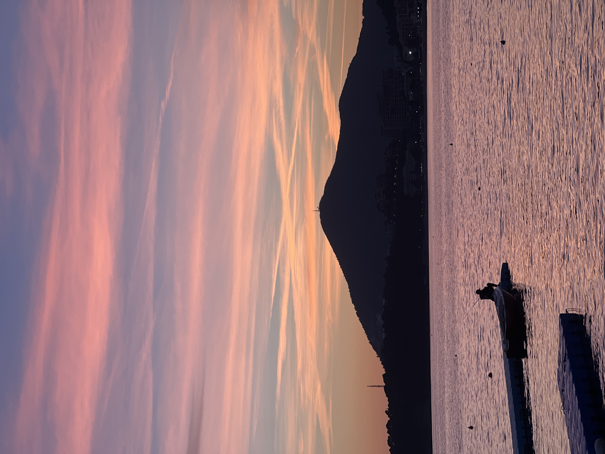 The image displays a coastal scene at dusk in Rafailovići, Montenegro. The sky, occupying the upper half of the frame, transitions from deep purple at the top to vibrant pink and orange hues in the middle, displaying numerous thin, horizontal cloud streaks. A large, dark mountain is silhouetted against the colorful sky, dominating the background horizon. A communication tower is clearly visible at the mountain's peak, and another slender, dark structure stands on a lower ridge to the left. Faint, scattered lights from coastal buildings are discernible along the mountain's base. The lower half of the image features a body of water, likely the Adriatic Sea, reflecting the sky's purple and pink tones with noticeable surface ripples. Several dark buoys are dispersed across the water. In the foreground, a modular floating dock, constructed from blue or grey plastic sections, extends into the scene from the left. A small, dark boat with a reddish hull is moored alongside this dock. One individual is seated in the boat, facing away from the viewer, silhouetted against the water, holding a fishing rod extended towards the right.