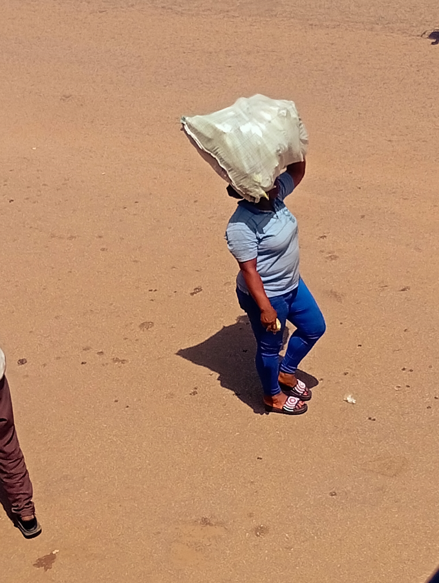 A woman walks along a dusty, light brown dirt road, carrying a large, light-colored sack of goods on her head. She is wearing a blue t-shirt, blue leggings, and patterned sandals. Her shadow is cast behind her. In the lower left of the frame, the legs and feet of another person wearing dark brown pants and dark shoes are visible. The background is an indistinct, flat, sandy or dirt surface, suggesting an outdoor environment, possibly on the outskirts of a town or city. The overall impression is of a common, everyday scene of a person engaged in the transport of goods. The location is identified as Bukuru, Nigeria.