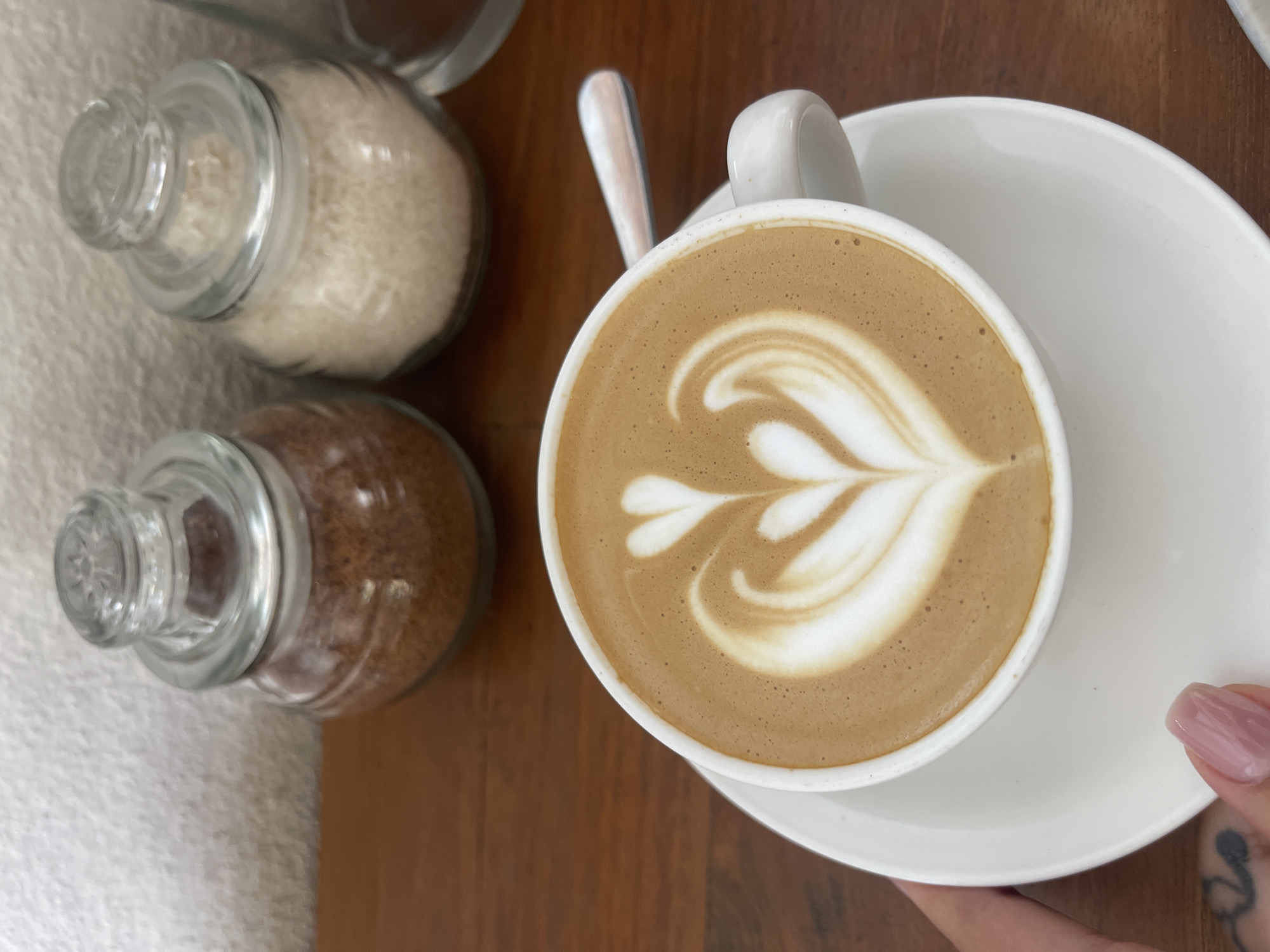In North Kuta, Indonesia, a white ceramic cup of latte art coffee sits on a white saucer on a wooden table. The latte art is a delicate white design on top of the brown coffee. To the left of the cup, a hand with manicured nails is visible, holding the saucer. In the background, two glass jars containing sugar are placed on the wooden table. The jar on the left contains brown sugar, and the jar on the right contains white sugar. A spoon rests on the saucer to the right of the cup. The background wall is white and textured.