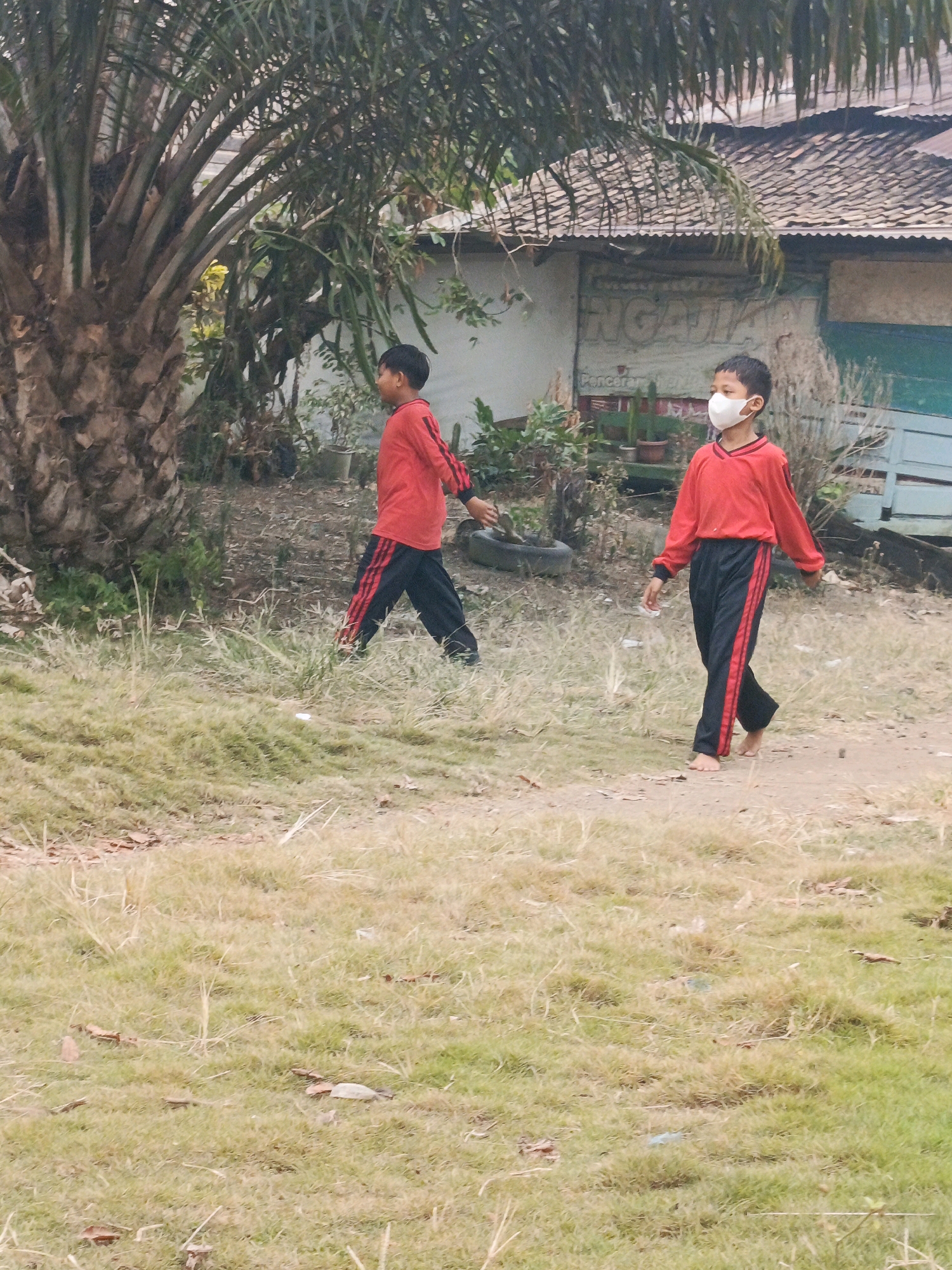 Two young boys, dressed in red long-sleeved shirts with black pants featuring red stripes down the sides, are walking on a dirt path in what appears to be a semi-rural area. The boy on the right is wearing a white face mask. Both boys are barefoot. In the background, a building with a corrugated metal roof and a sign with Indonesian writing is visible, along with various plants and a palm tree. The ground is covered with dry grass and dirt. The scene suggests a warm climate, possibly in the early morning or late afternoon given the lighting. The location is identified as Megang Sakti II, Indonesia.