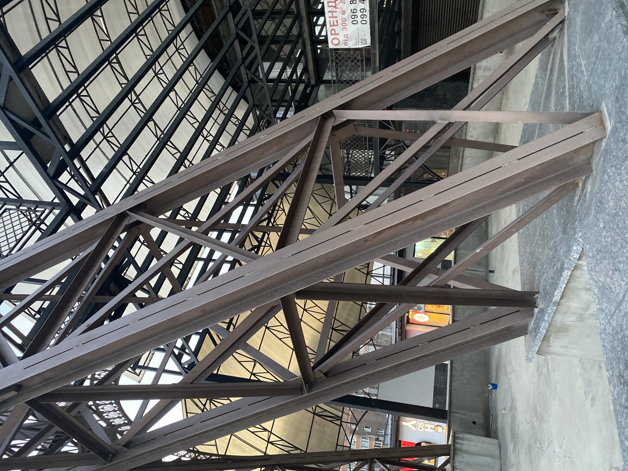 The image depicts a close-up view of a large, weathered steel truss structure, likely part of an outdoor market or covered walkway, in Vinnytsia, Ukraine. The structure is composed of diagonal and horizontal beams, showing signs of rust and age. Above, a translucent white canopy is supported by a dark metal framework. In the background, a building with storefronts is visible, including one with neon signage. A white sign with red and black text, indicating "ORENDA" (rent in Ukrainian) and phone numbers, is affixed to the building. The ground appears to be paved with concrete or stone. A small blue plastic container is present on the ground in the lower left quadrant of the image. No people are clearly discernible in the scene.