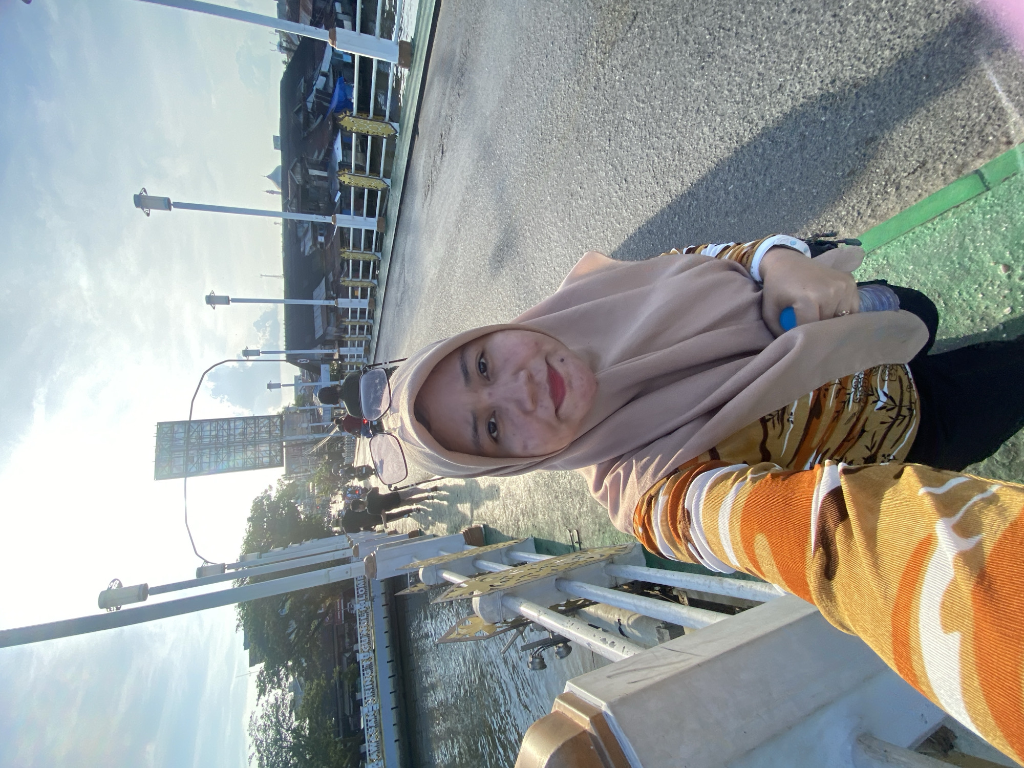 A woman wearing a beige hijab and a long-sleeved shirt with an orange, white, and brown striped pattern is taking a selfie on a bridge. She is holding a blue water bottle in her right hand. She is standing on a paved road on the bridge, with a body of water to her left. In the background, there are buildings with tin roofs, trees, streetlights, and several people on motorcycles. The scene appears to be in Banjarmasin, Indonesia, based on the architecture and the presence of canals. The sun is shining brightly, casting shadows on the bridge.