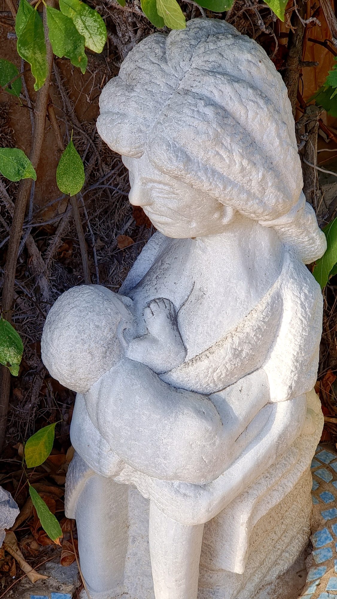 This is a medium shot of a white stone sculpture of a mother breastfeeding a baby. The sculpture is outdoors, with dried branches and green leaves in the background. The mother's hair is styled in a bun, and she is looking down at the baby. The baby is partially covered by a cloth. In the bottom right corner, a small mosaic of blue tiles is visible. The sculpture is likely located in a garden or public space in Sant Joan les Fonts, Spain, given the context.