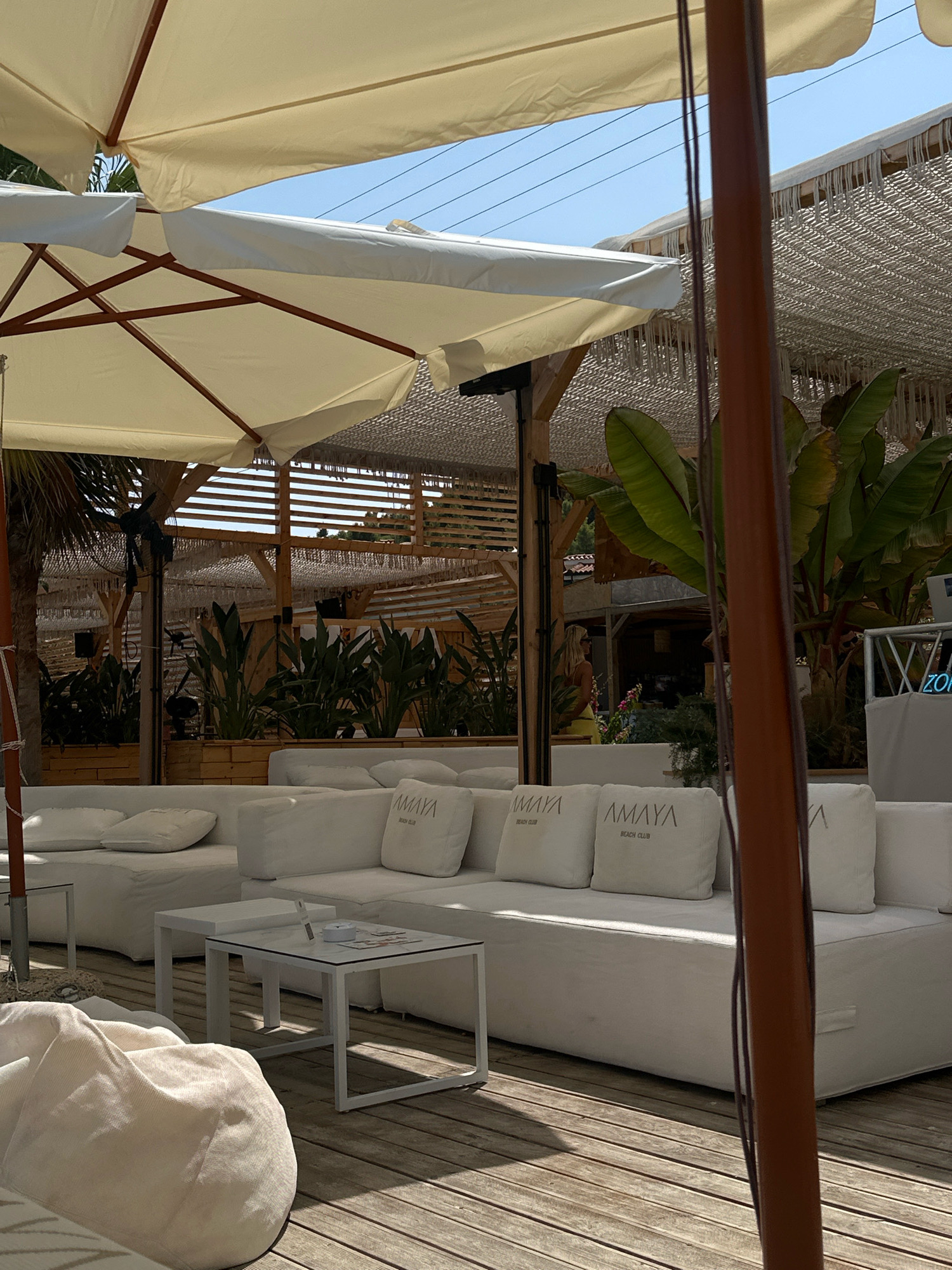 The image depicts an outdoor seating area at what appears to be a beach club in Pallini Municipal Unit, Greece. The foreground features a large, white modular sofa with several white cushions, some of which have "AMAYA BEACH CLUB" branding. In front of the sofa, a small white square coffee table is visible, with an ashtray and some magazines on it. To the left of the sofa, a cream-colored beanbag chair is partially visible. The seating area is set on a wooden deck. Overhead, large cream-colored umbrellas provide shade, supported by wooden poles. In the background, there is more seating, planters with lush green foliage, and a structure with a thatched roof and horizontal wooden slats. A person, a woman with blonde hair wearing a yellow top, is visible in the background walking away from the camera. The sky is clear and blue.
