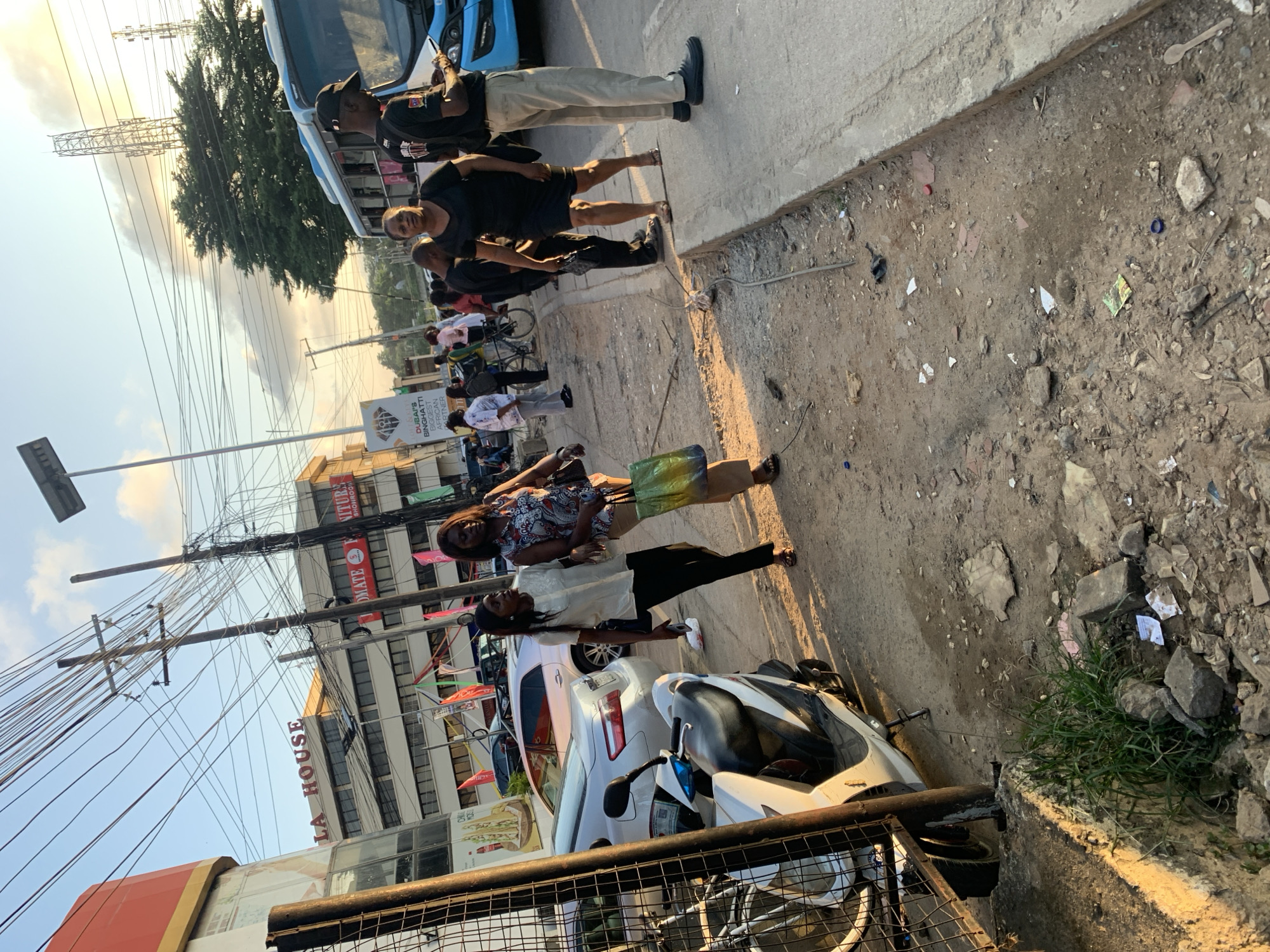 The image depicts a street scene in Ikeja, Nigeria, with a focus on a sidewalk area cluttered with debris and a nearby road. In the foreground, a white scooter is parked next to a metal fence. Two women are walking on the sidewalk, one carrying a green shopping bag. Behind them, a blue bus is visible, suggesting a transportation hub or a busy street. Several cars are parked on the road. The background shows buildings, including one with "LA HOUSE" signage, and power lines crisscrossing the sky. The overall environment appears to be a commercial or urban area with signs of everyday activity and some disarray.