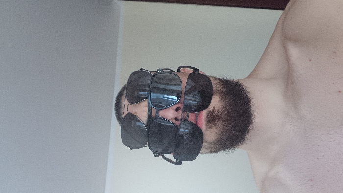 An adult male is depicted indoors in Sofia, Bulgaria, shown from the upper chest upwards. He has short hair and a dark beard, and his upper torso and shoulders are bare. His face is largely obscured by four distinct pairs of sunglasses, vertically stacked. The top pair features silver-toned aviator-style frames with dark lenses. Immediately below, a pair with dark, rectangular frames and dark lenses is positioned. A third pair, with dark frames and dark lenses, rests across the bridge of the nose and partially on the upper lip. The lowest pair, also dark-framed and dark-lensed, is positioned across the mouth and supported by the chin. Reflections of interior light sources are visible in the lenses of the sunglasses. The background consists of a plain, light beige wall, with a light gray ceiling visible above. A dark brown vertical element, consistent with a door frame or similar structure, is present on the far right.