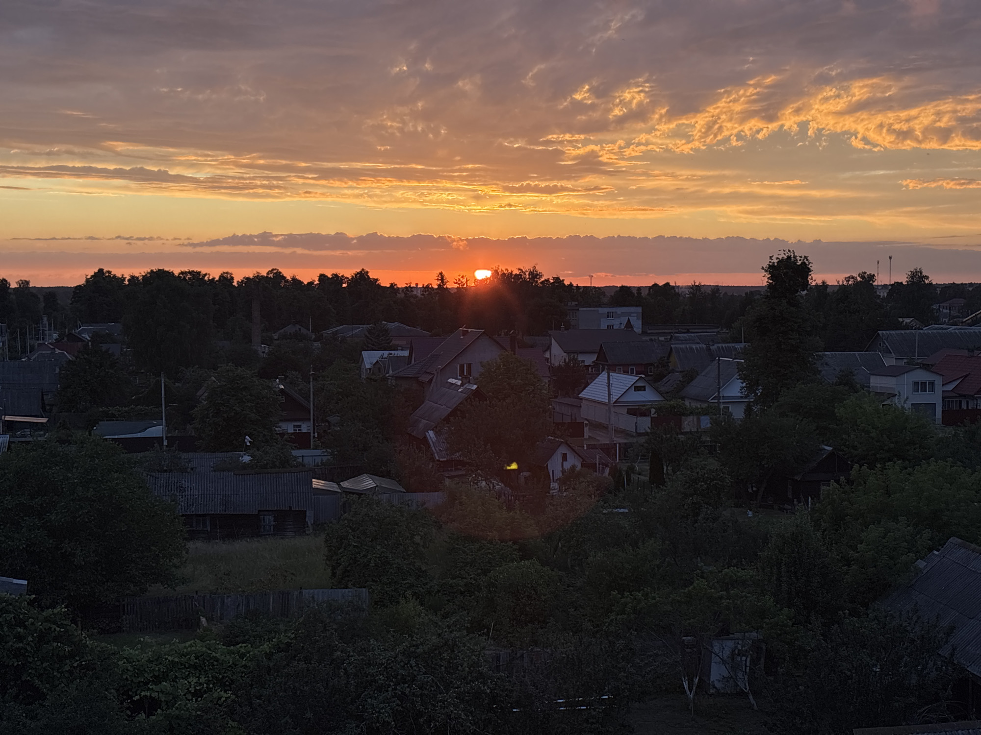 An aerial, high-angle, full shot captures a sunset over a rural village in Pratasevichi rural council, Belarus. The sky is a dramatic gradient of orange, yellow, and pink, with wispy clouds catching the last rays of sunlight. Below, the village is silhouetted against the vibrant sky, with the dark shapes of trees and rooftops filling the lower half of the frame. The village appears to be sparsely populated, with houses of varying sizes and styles scattered among the trees. The overall impression is one of peaceful serenity and the natural beauty of the Belarusian countryside at dusk.