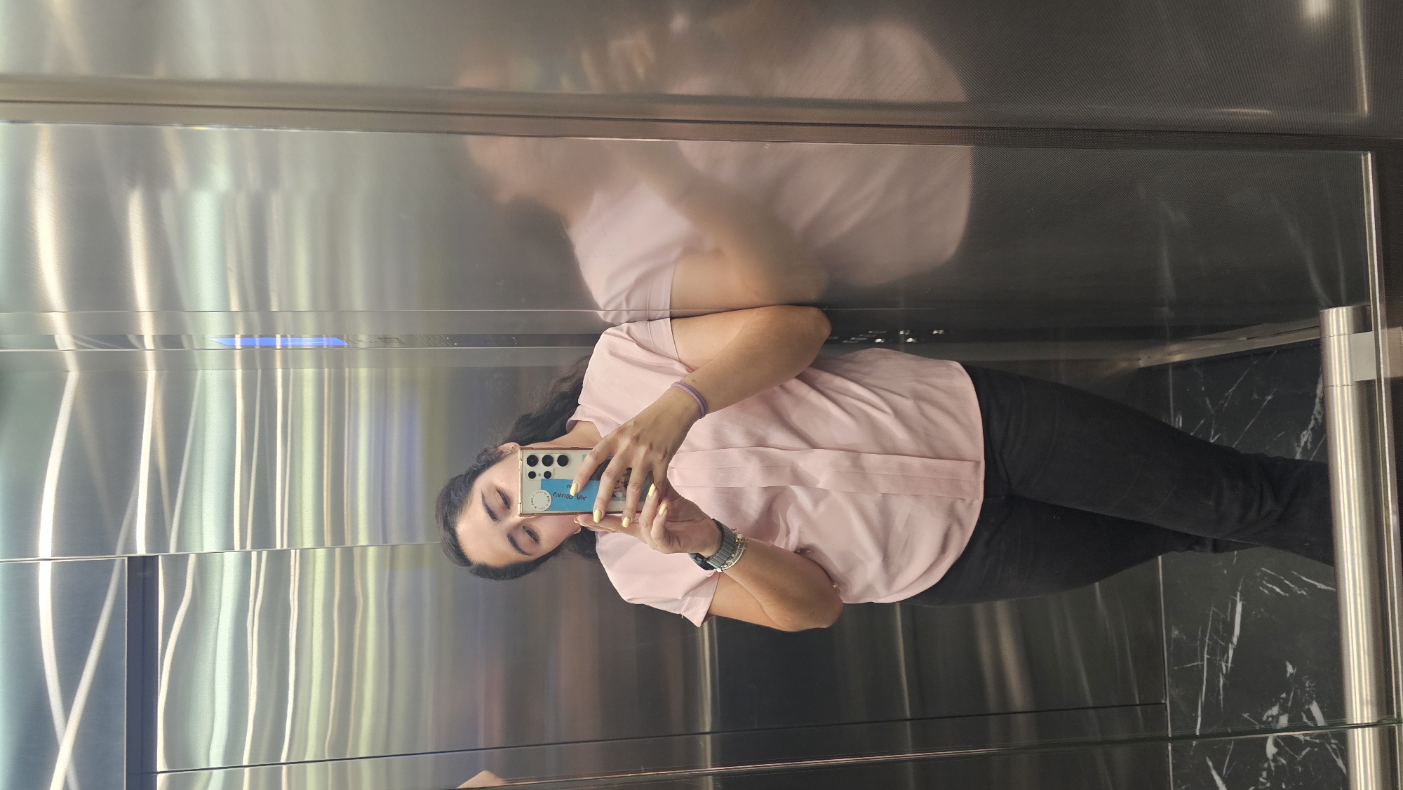 A young woman stands inside an elevator, taking a selfie with her smartphone. She wears a light pink, short-sleeved shirt and dark pants. Her dark hair is pulled back, and she's wearing a bracelet on her left wrist. The elevator interior features stainless steel walls and a dark marble-patterned floor. The city of Puebla, Mexico, is not visually confirmed in this image.