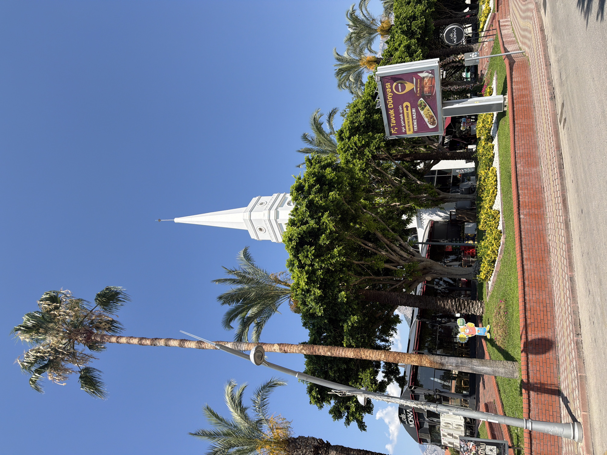 The image shows a street view in Kemer, Turkey, on a clear, sunny day. In the foreground, a paved sidewalk and road are visible, bordered by a low brick wall. A streetlamp stands on the left side of the image. Behind the wall, a lush green lawn is dotted with yellow flowers. Several palm trees are scattered throughout the scene, with one tall, mature palm tree dominating the left side of the frame. Behind the trees, a modern building with a black facade and large glass windows can be seen, featuring a sign that reads "Cafe MOKKA". Further back, a white building with a tall, pointed spire, resembling a church steeple, rises against the blue sky. To the right of the image, a large billboard advertises "Tavuk Dünyası," a Turkish restaurant chain, displaying images of food and a map pin. The overall impression is of a vibrant, Mediterranean urban environment with a mix of commercial buildings, greenery, and a distinct architectural element.