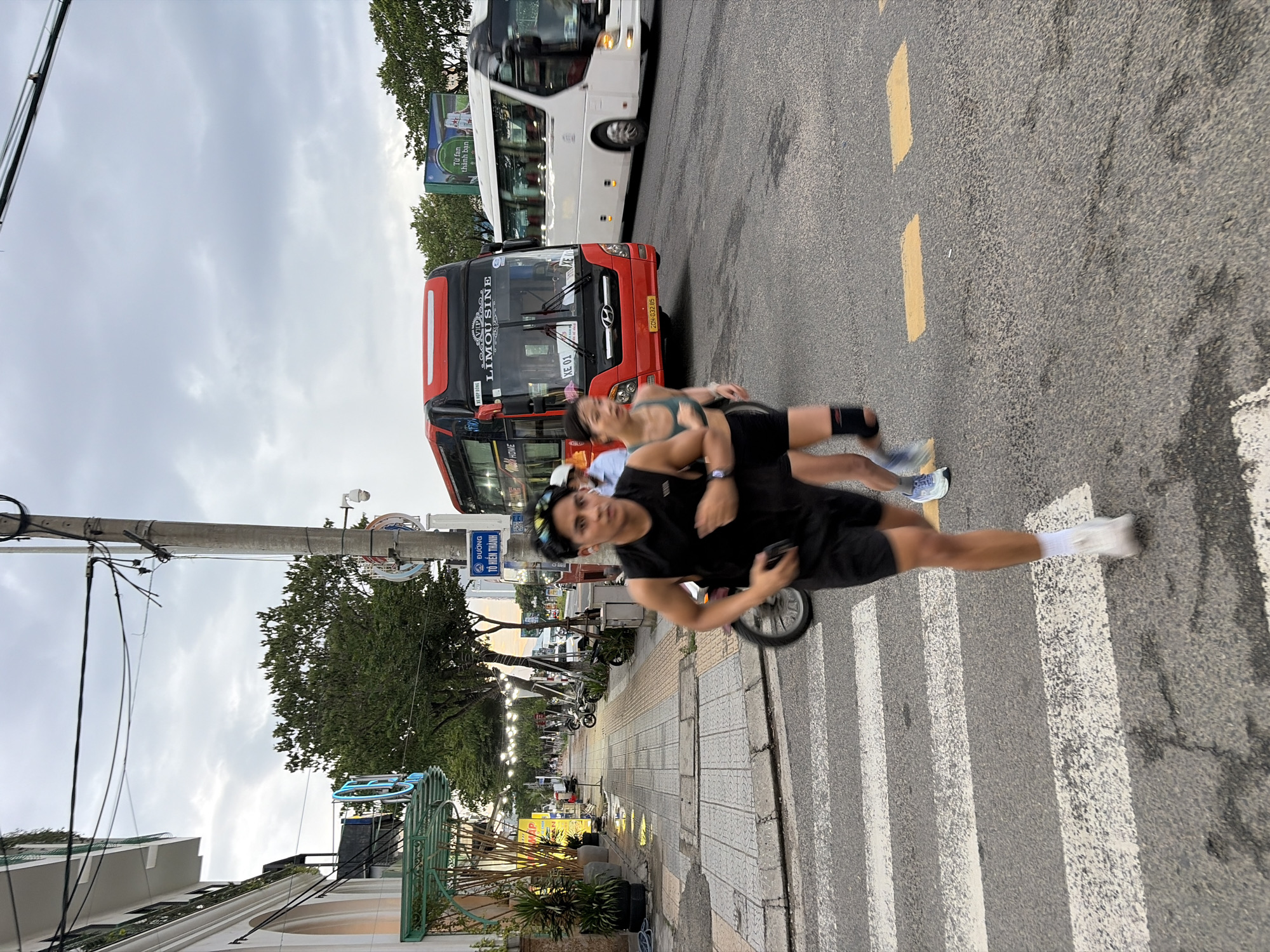 The image depicts a street scene in Da Nang, Vietnam, likely during the day under a cloudy sky. In the foreground, two individuals are running across a crosswalk. The person on the left is a male wearing a black sleeveless shirt, black shorts, white socks, and white running shoes, with sunglasses on his head. The person to his right is a female wearing a green athletic top and black shorts, with knee sleeves on her legs. Behind them, a red limousine bus is stopped or moving slowly in the street, with the text "LIMOUSINE" visible on its front. To the right of the limousine bus, a white passenger bus is in motion. The background shows a sidewalk with trees, buildings, and streetlights. A utility pole with wires is also prominently visible on the right side of the image. The overall environment suggests a commercial or busy urban area.